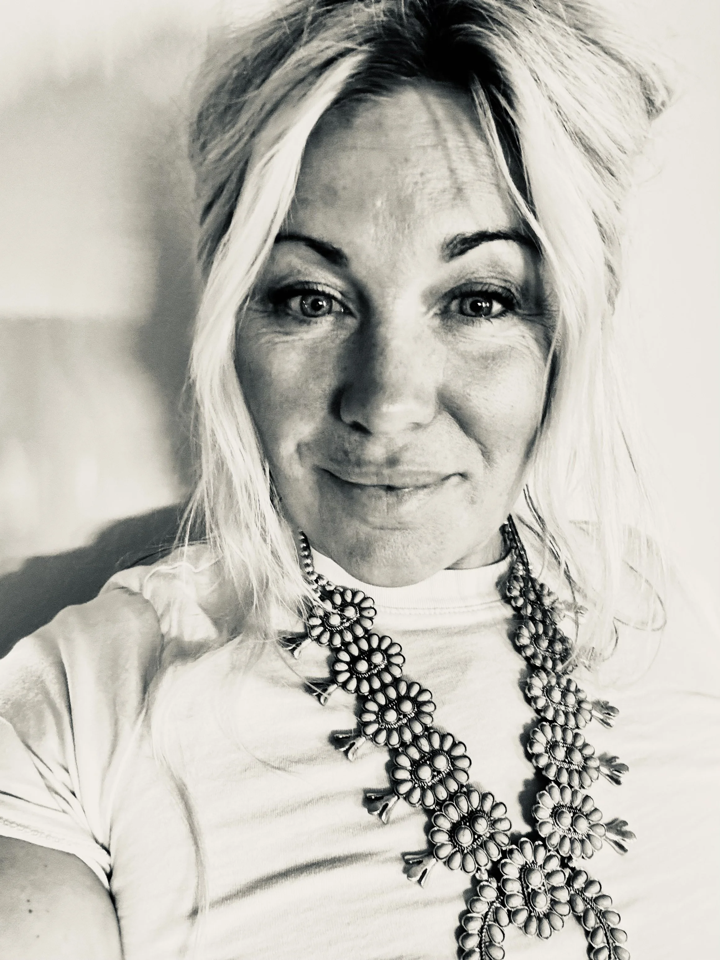 Black and white close-up photo of a woman with wavy blond hair, wearing a necklace with large floral-shaped beads and a light-colored shirt, smiling slightly.