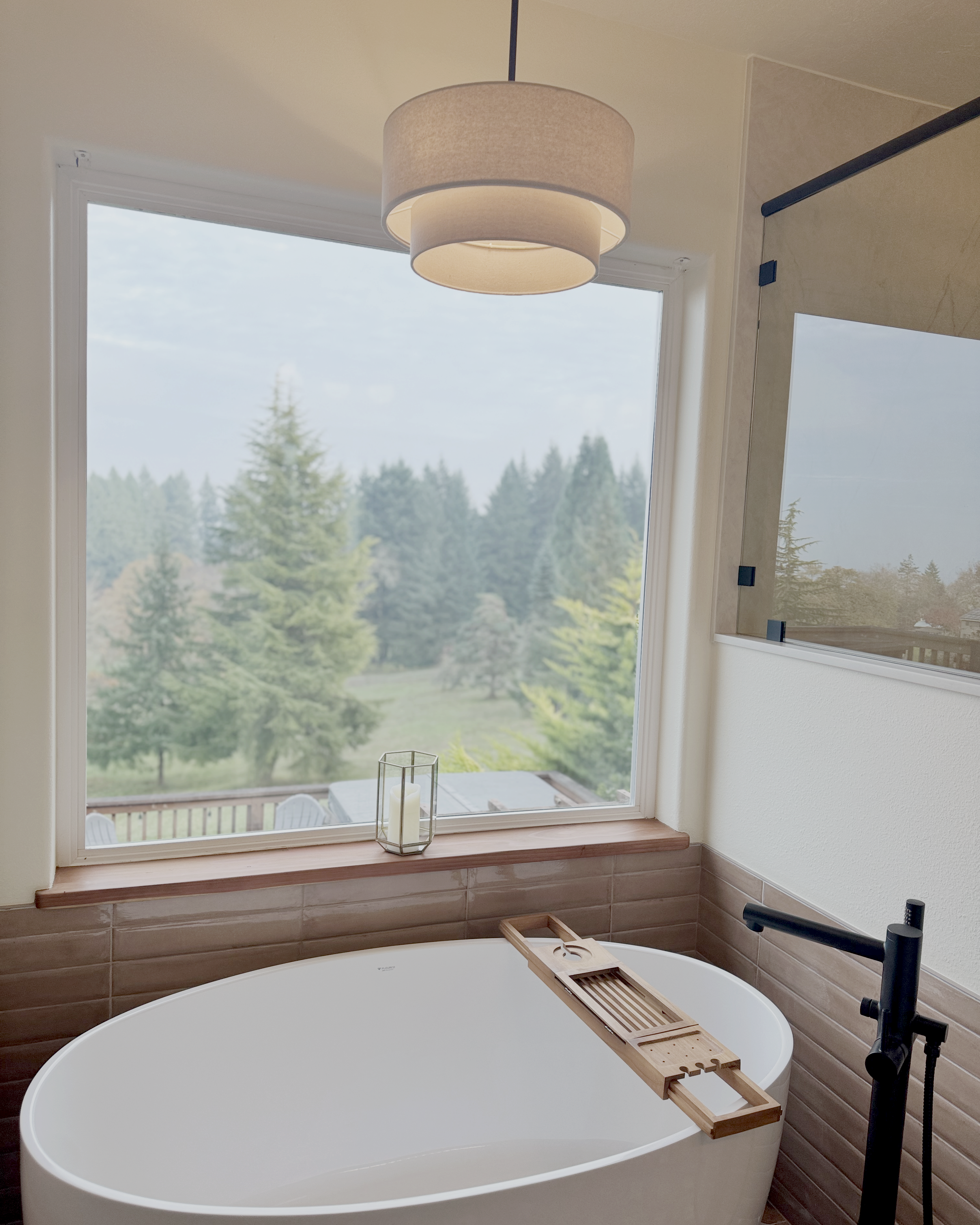Modern bathroom with a white freestanding bathtub, a large window with a scenic view of trees, a wooden tray on the bathtub, a candle in a glass holder on the window ledge, a ceiling light fixture, and a black floor-mounted faucet.