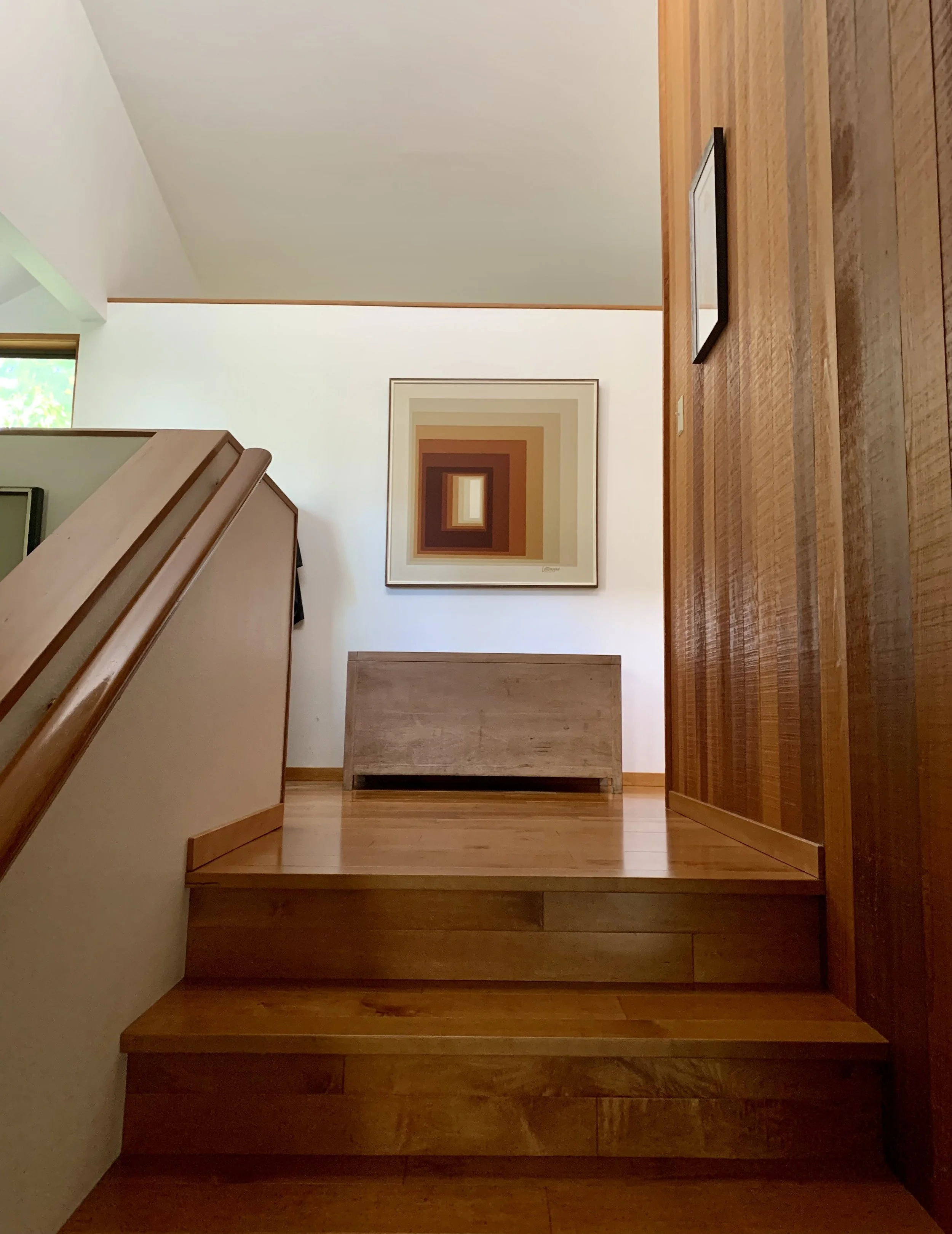 Wooden staircase leading up to a landing with a wooden bench, framed artwork with concentric rectangles in shades of brown and orange, and a piece of framed art on the wall.