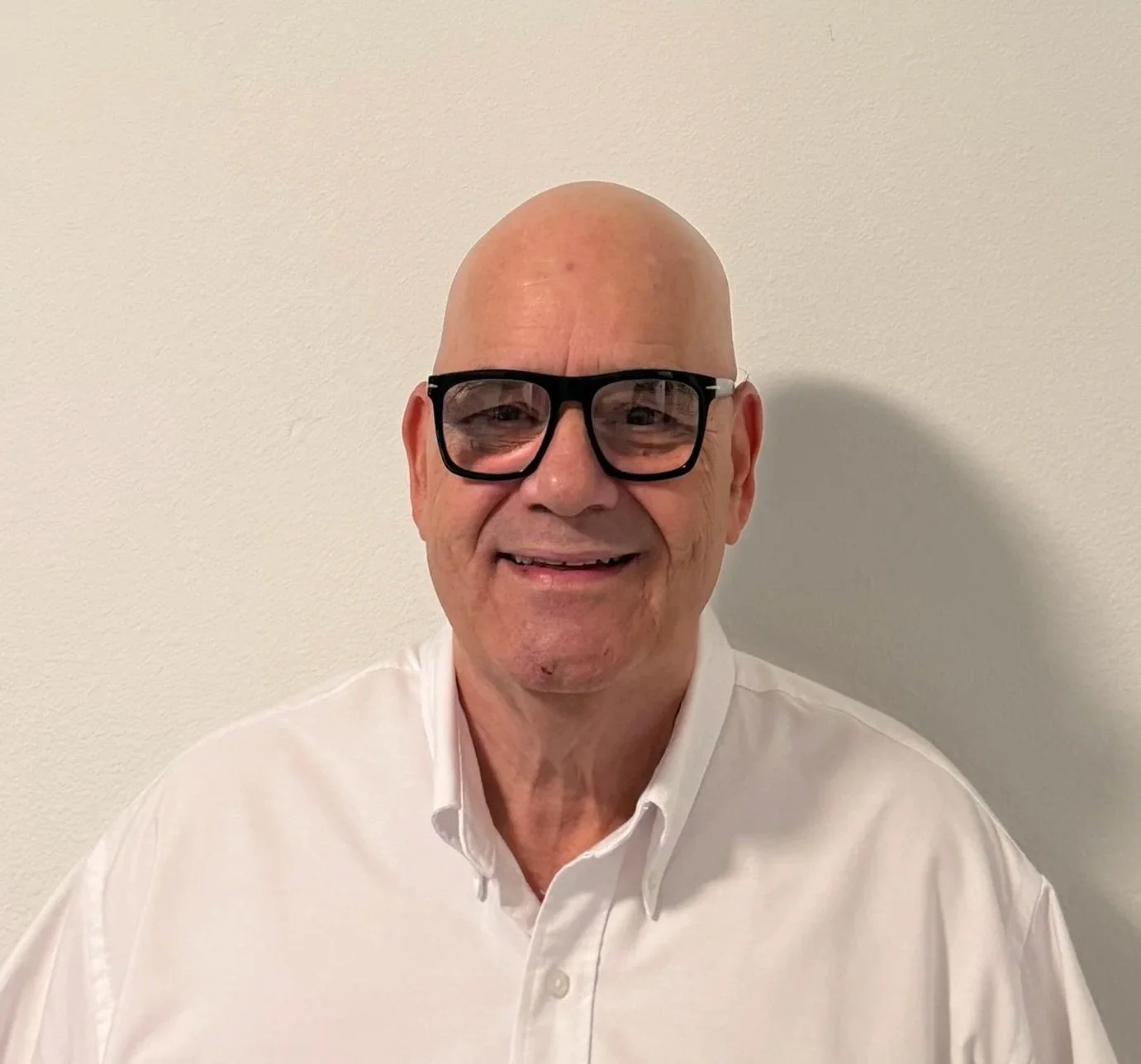 Smiling man with glasses and a bald head, wearing a white dress shirt, standing against a plain white wall.