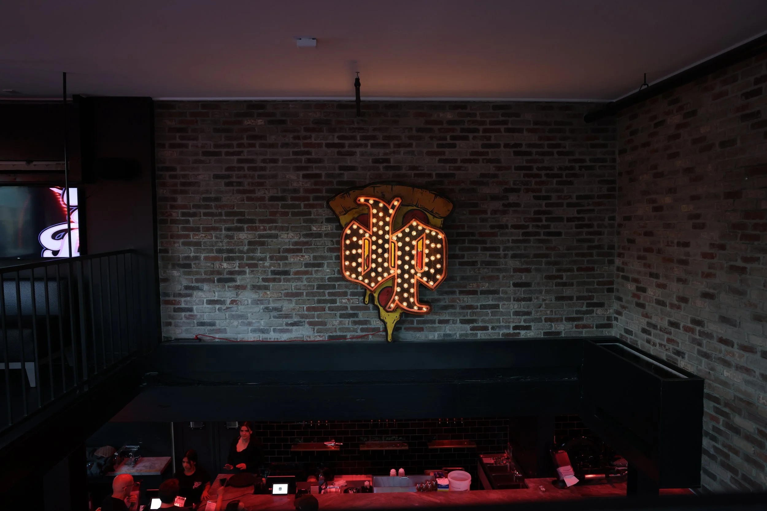Interior of a bar with a neon sign on a brick wall, and people working behind the bar.