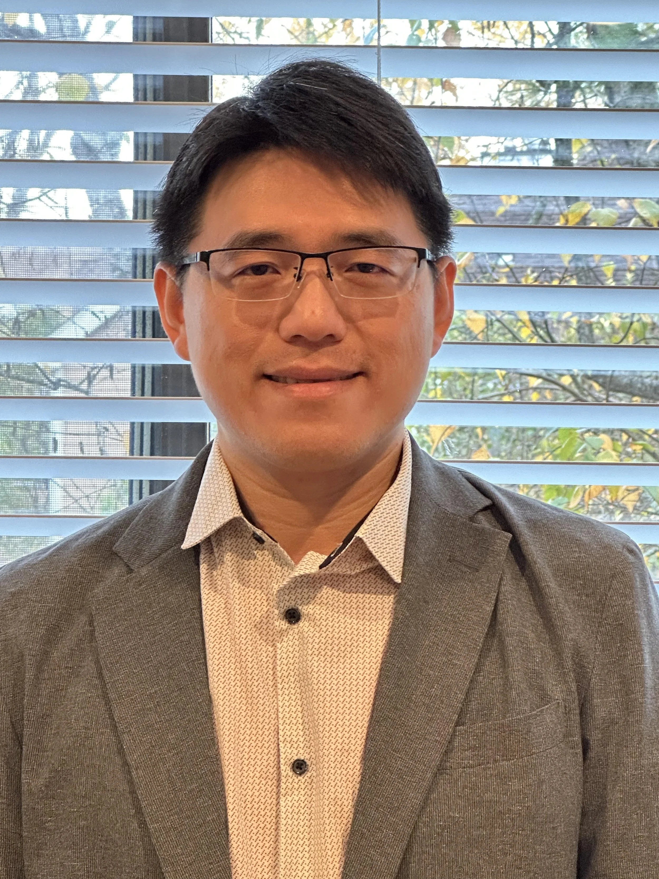 A man wearing glasses, a white shirt, and a dark blazer standing indoors with large windows in the background.
