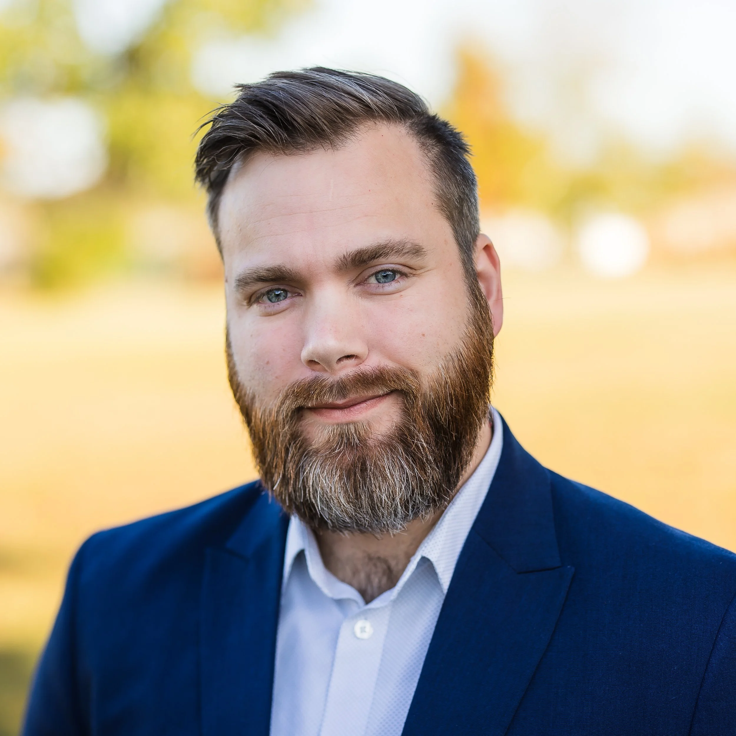 A portrait of a man with a beard and short dark hair, wearing a dark blue suit jacket and white shirt, standing outdoors with blurred trees and grass in the background.