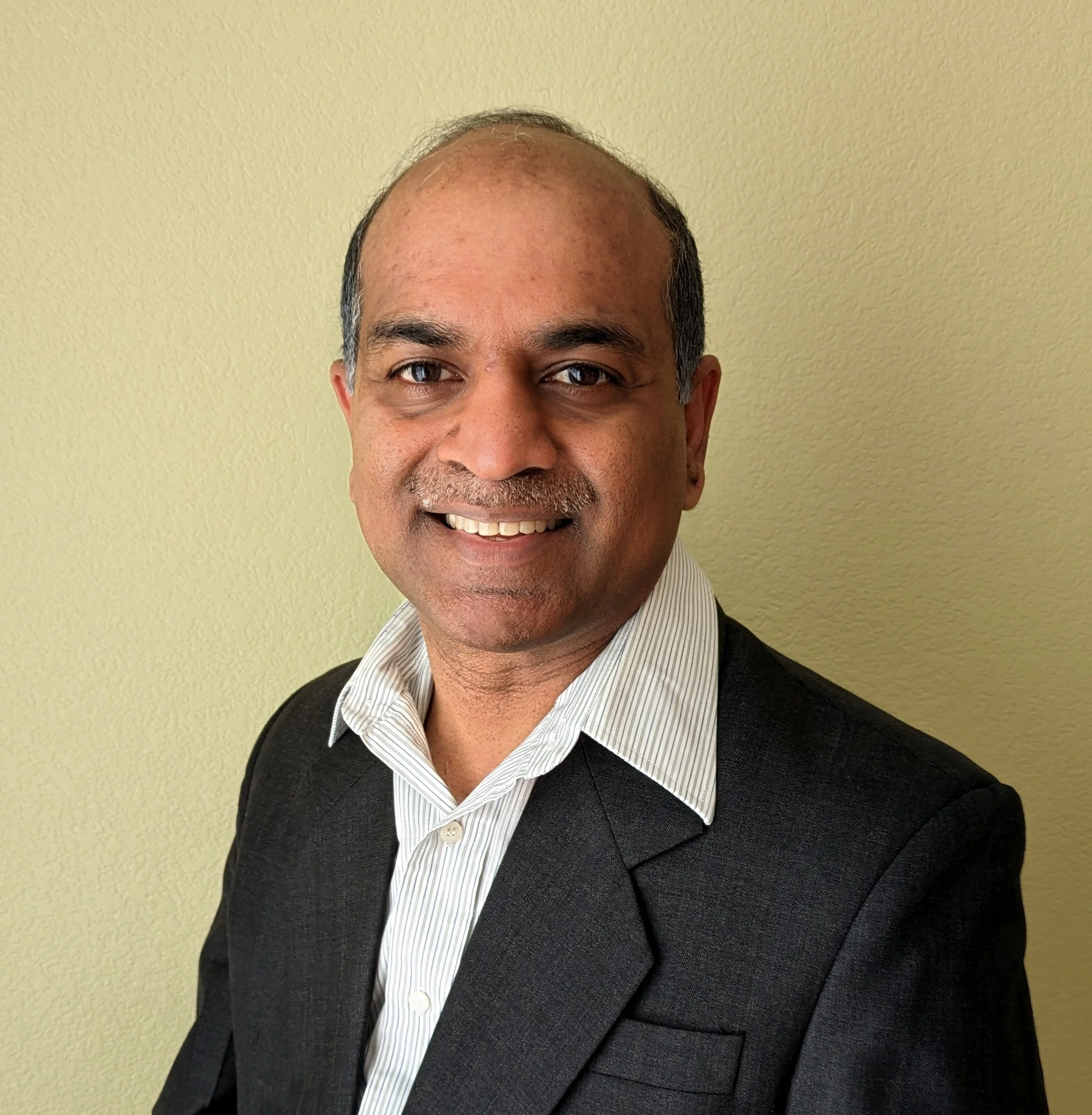 A man with curly dark hair, glasses, and a beard, wearing a black sweater, looking thoughtfully to the side, with a light-colored wall and a plant in the background.