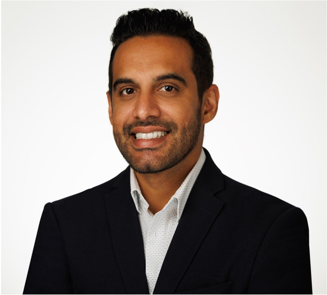 A man with curly dark hair, glasses, a nose ring, and a beard, looking thoughtfully to the side, in a room with a blurred plant in the background.