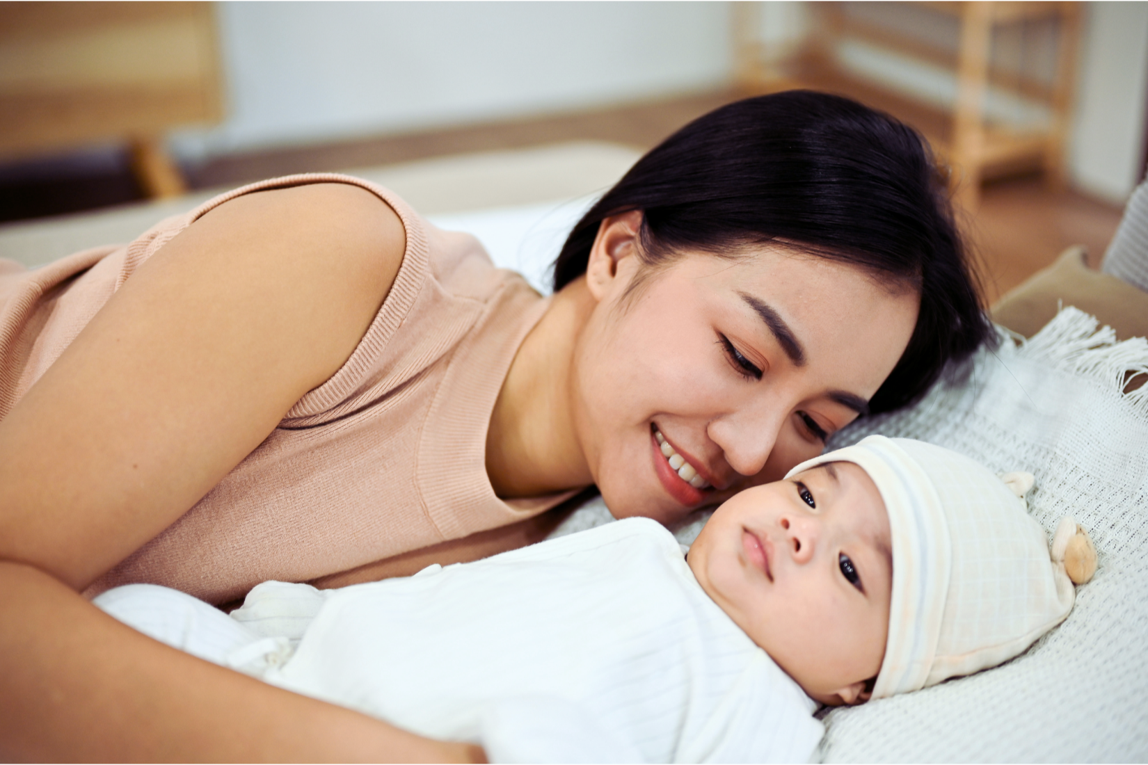 A woman lying on her side, smiling, looking at a baby lying on a pillow, in a cozy indoor setting.