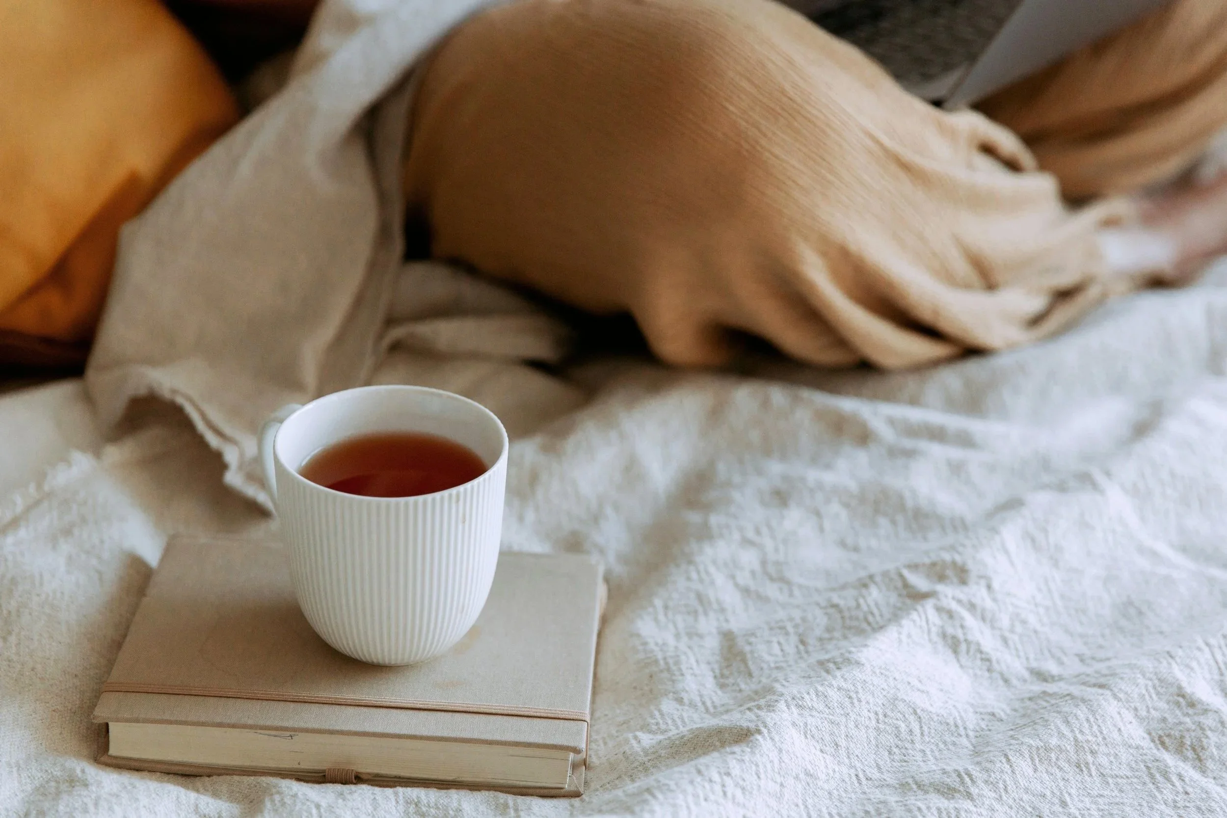 A white mug filled with tea resting on a book that is on a white, textured cloth surface, with beige and white fabric around.