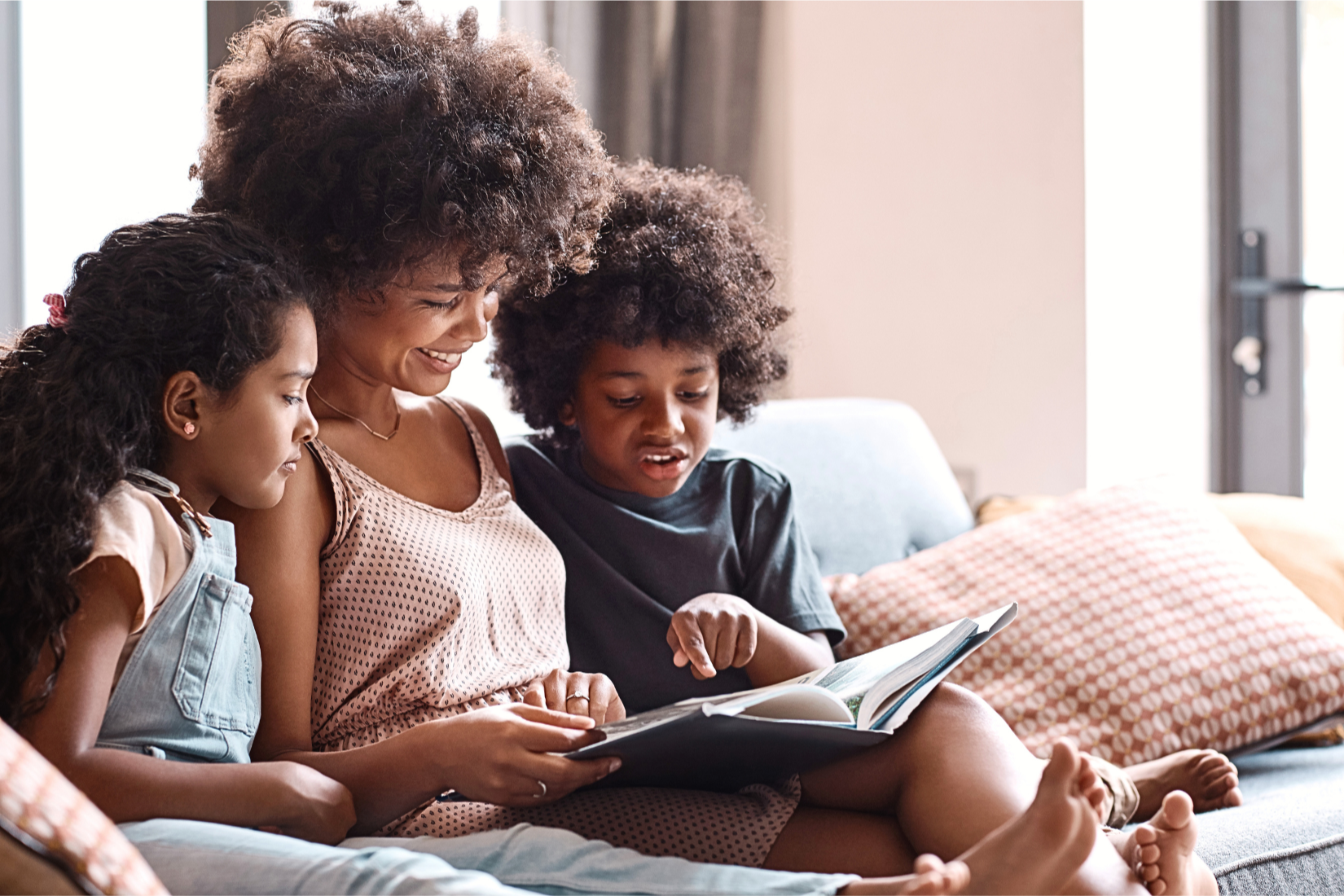 A smiling woman reading a book with two children, one boy and one girl, sitting on her lap on a couch. They are all looking at the book together.