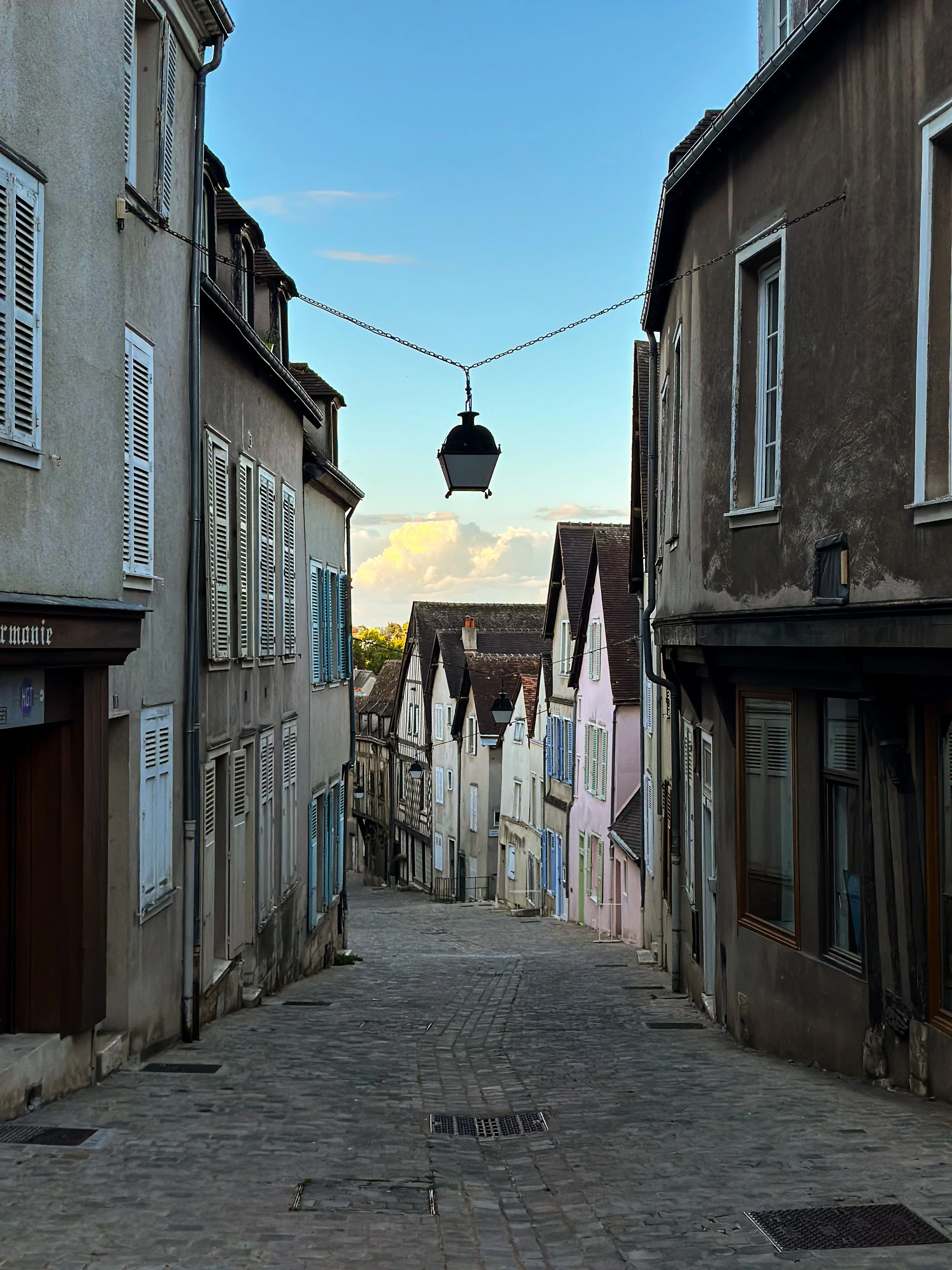 A narrow cobblestone street winding downhill between pastel-colored buildings with closed shutters on windows, a hanging lantern above, and a partly cloudy sky in the background.