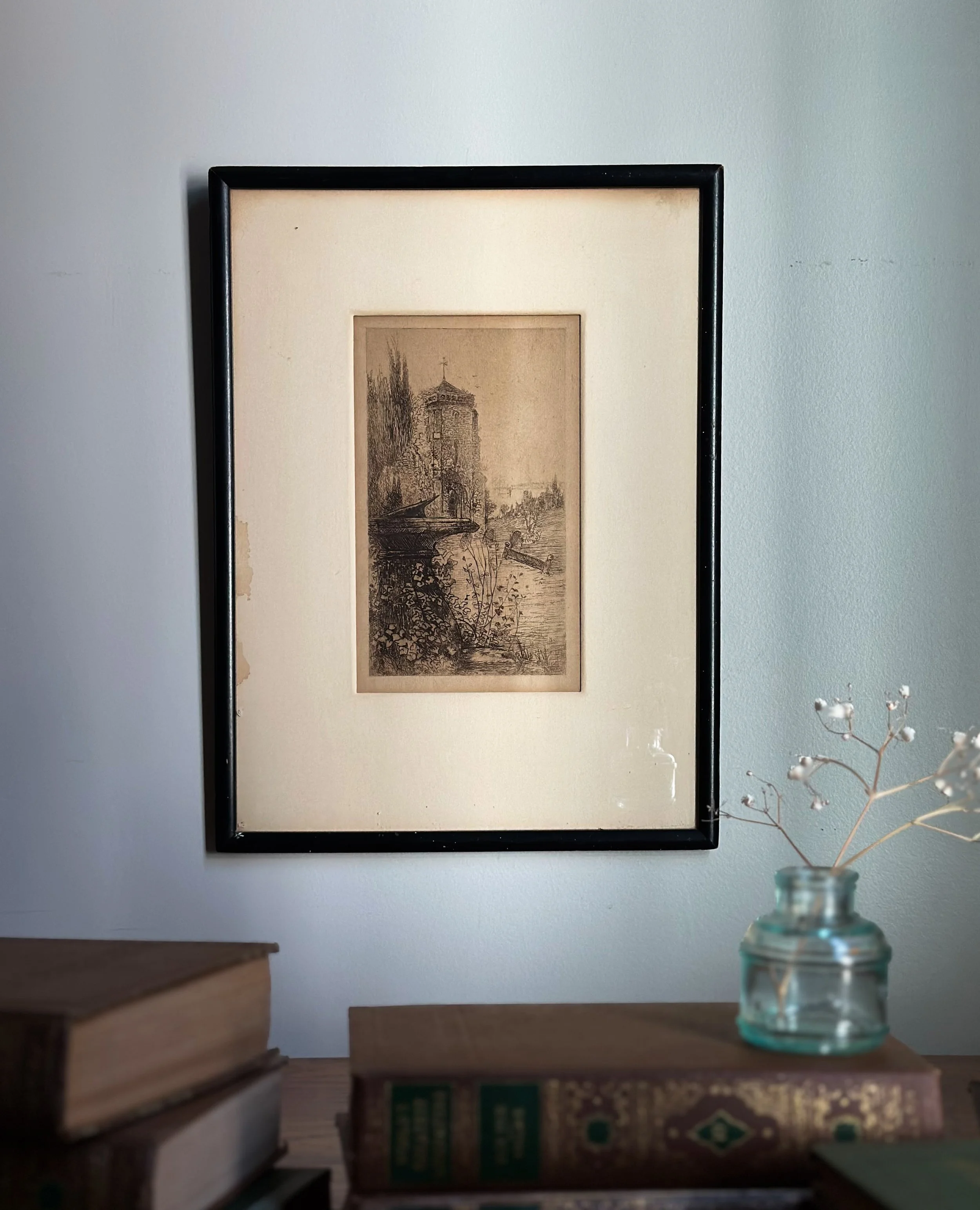 Framed vintage black and white sketch of a castle by a river, hung on a light-colored wall above a wooden shelf with books and a small glass jar with dried flowers.