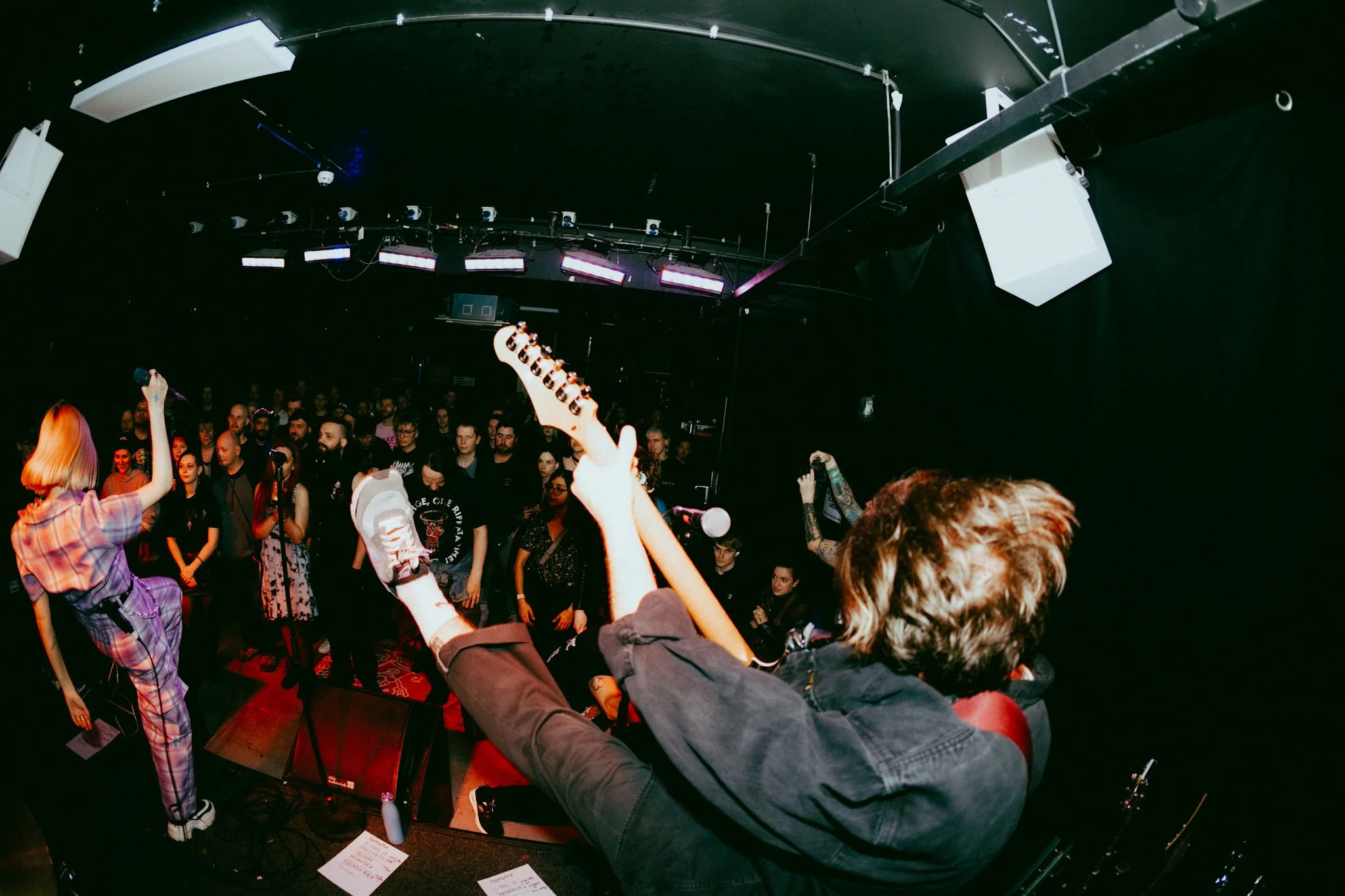 A live music performance with a female singer holding a microphone, a guitarist playing, and an audience watching in a dark venue with stage lights.