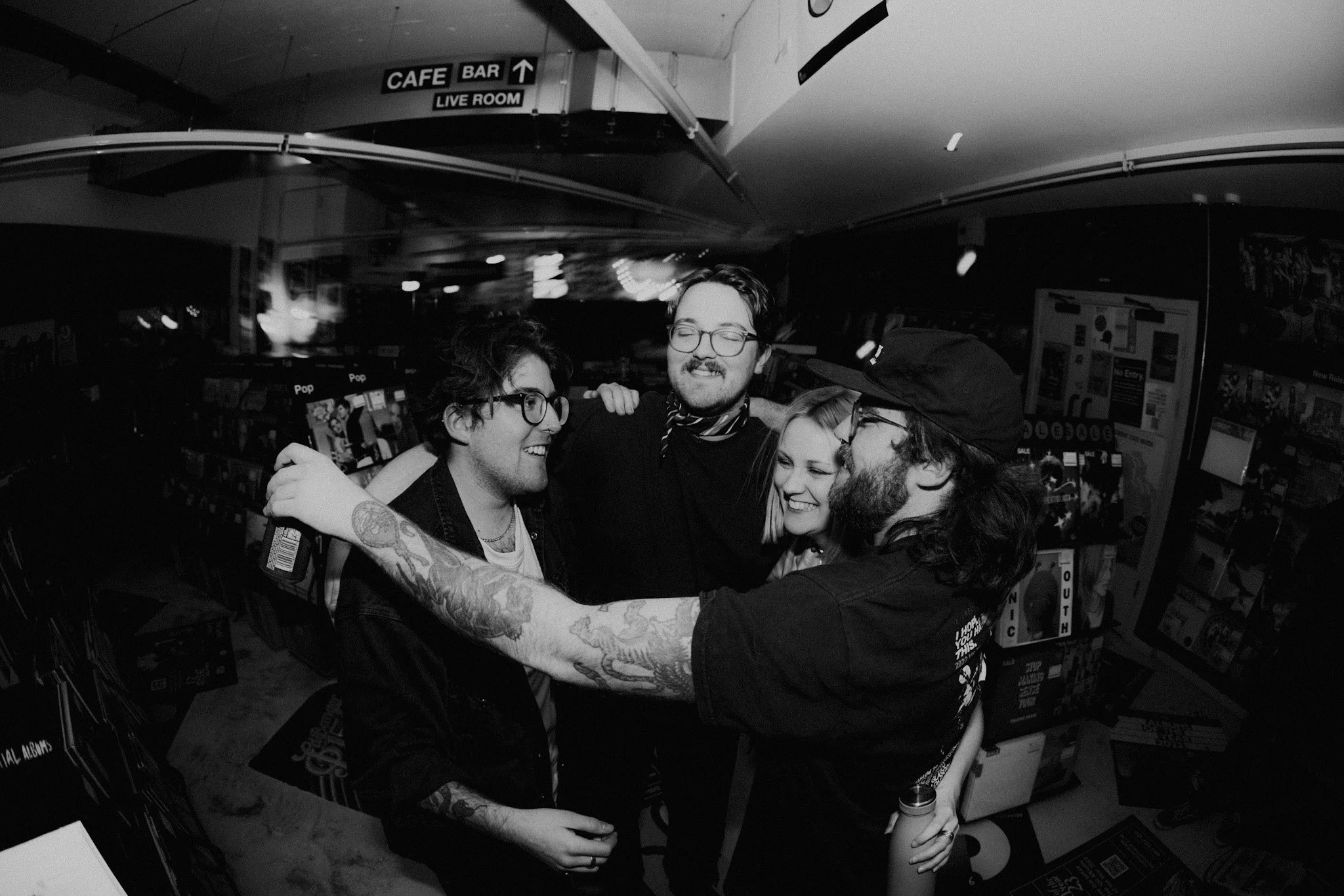 A group of four friends, three men and one woman, smiling and hugging each other in a store or record shop. The black and white photo shows them celebrating together.