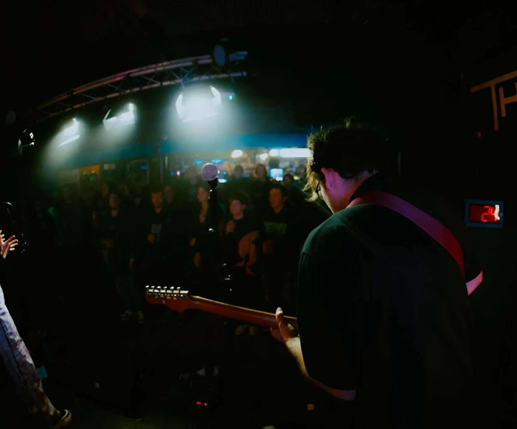 A musician playing an electric guitar on stage during a live concert with an audience watching, stage lights illuminating from above, and a digital clock showing 2:4.
