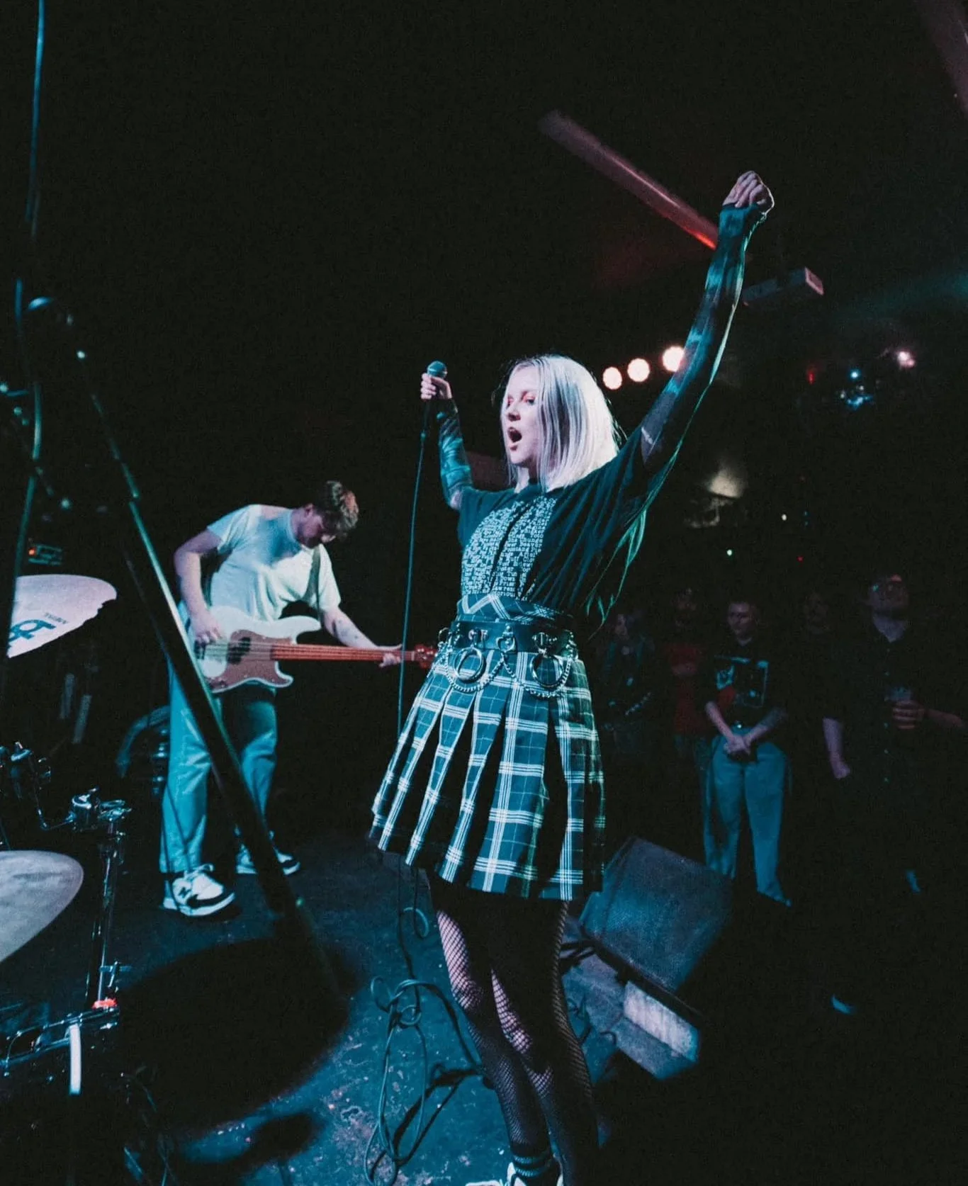 Female singer with platinum blonde hair performing on stage, holding a microphone in her right hand, with a guitarist in the background playing bass guitar, audience watching in the dark.