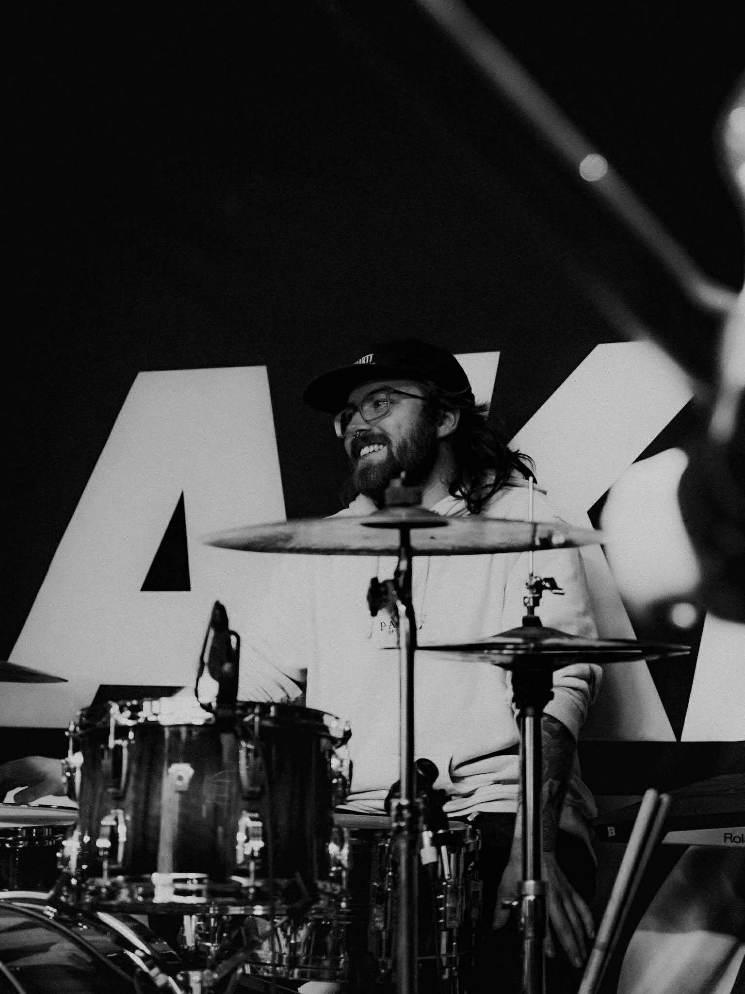 A man with glasses and a beard is playing drums, smiling, with large letters in the background.