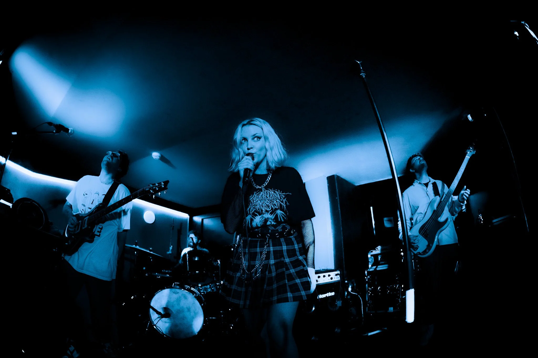 Female lead singer with blonde hair holding a microphone, performing with two male guitarists in a dimly lit venue.
