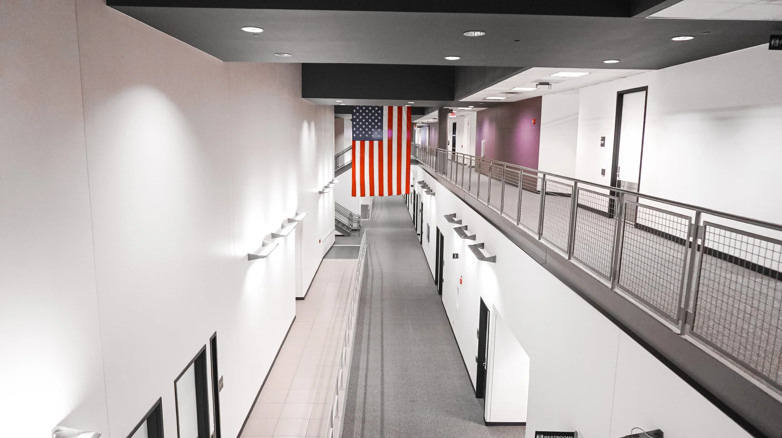 Empty indoor corridor with white walls and ceiling, gray carpeted floor, metal railing on the upper level, and an American flag hanging from the ceiling.