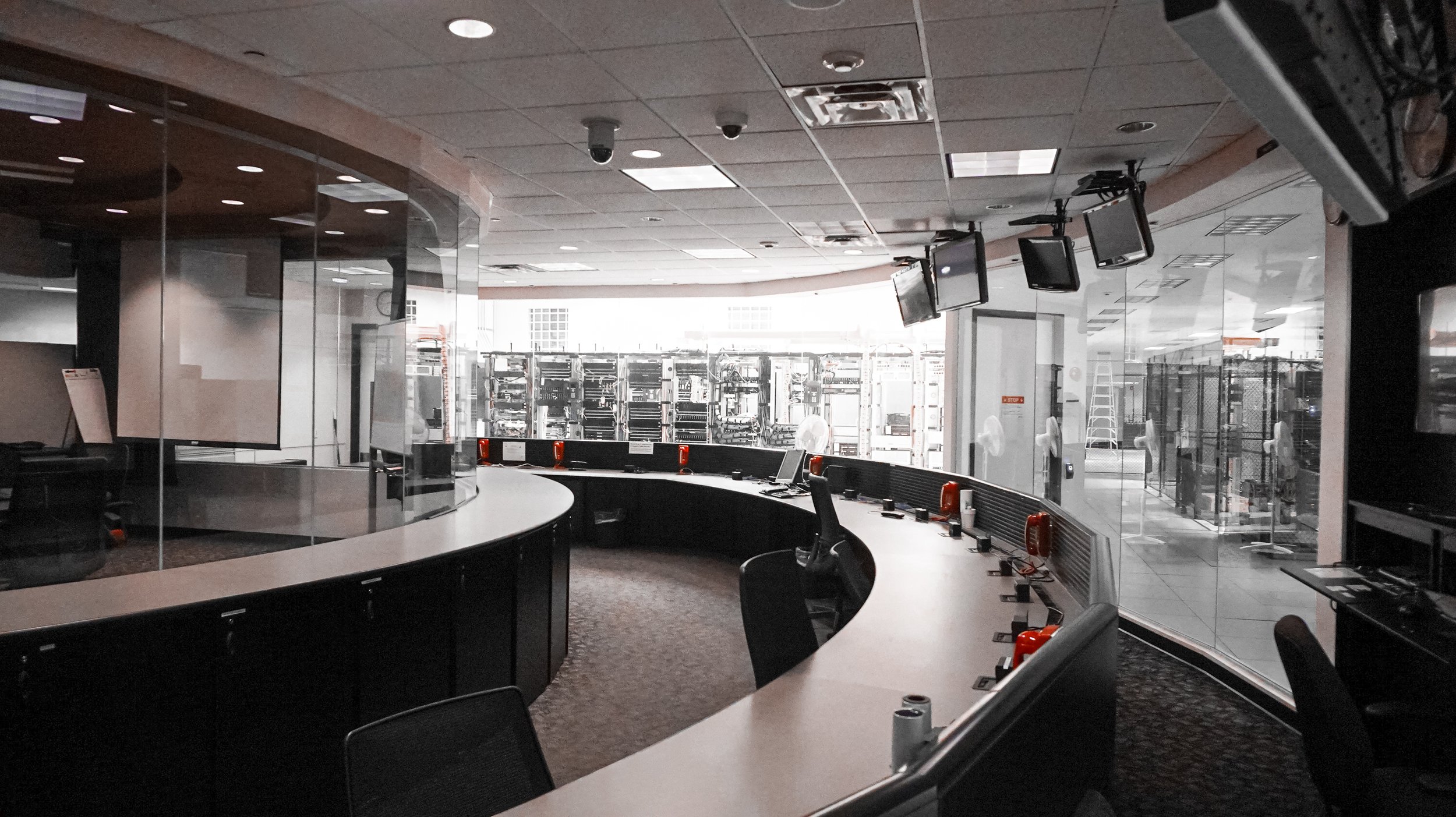 Empty control room or operations center with curved desk, multiple monitors hanging from ceiling, and server racks outside the glass wall.
