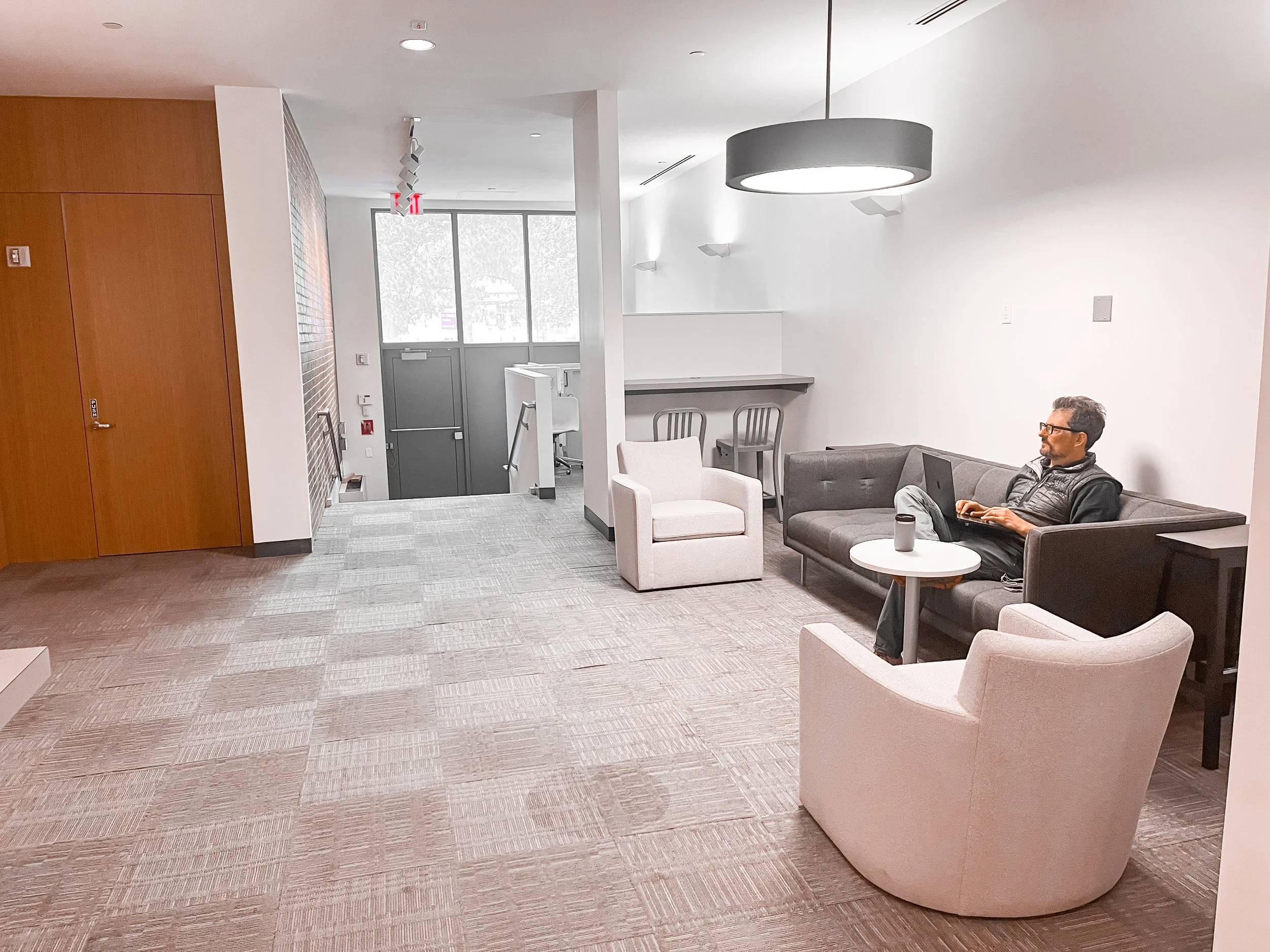 A man sitting on a gray sofa in a modern lounge area, working on his laptop, with a coffee mug on a small round table beside him. The room has beige carpet, a white wall, and minimal decor, with a large black circular hanging light and a beige armchair nearby.
