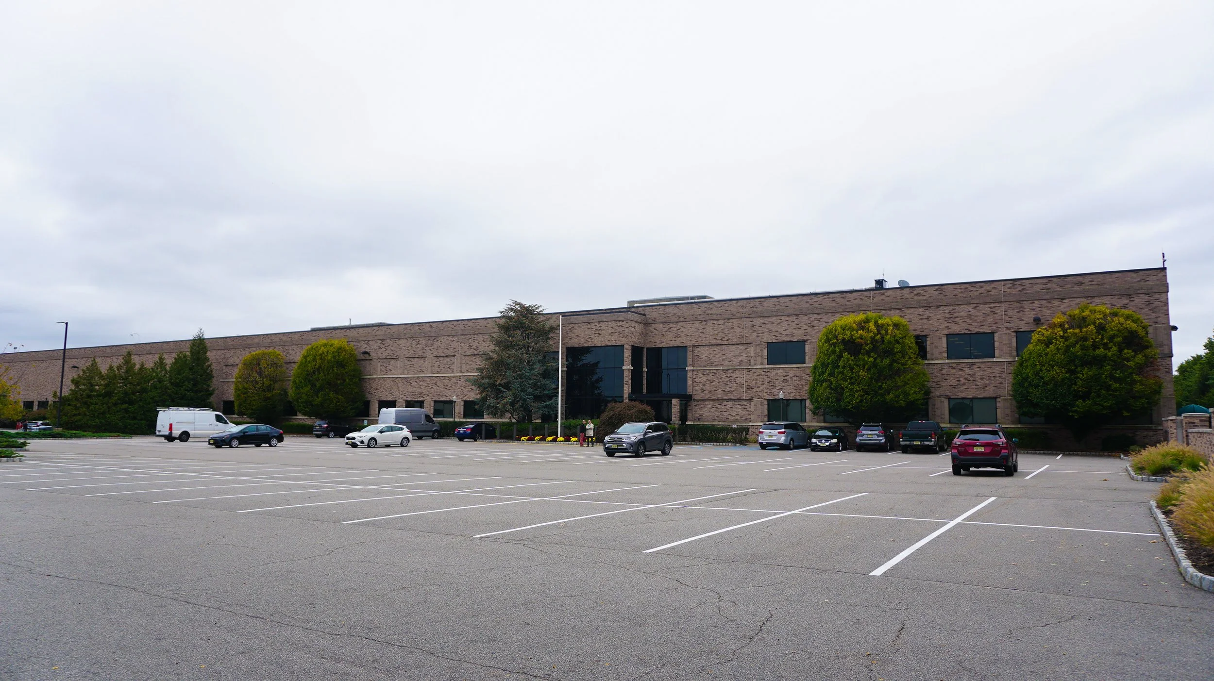 Large brick office building with several trees and parked cars in front, overcast sky