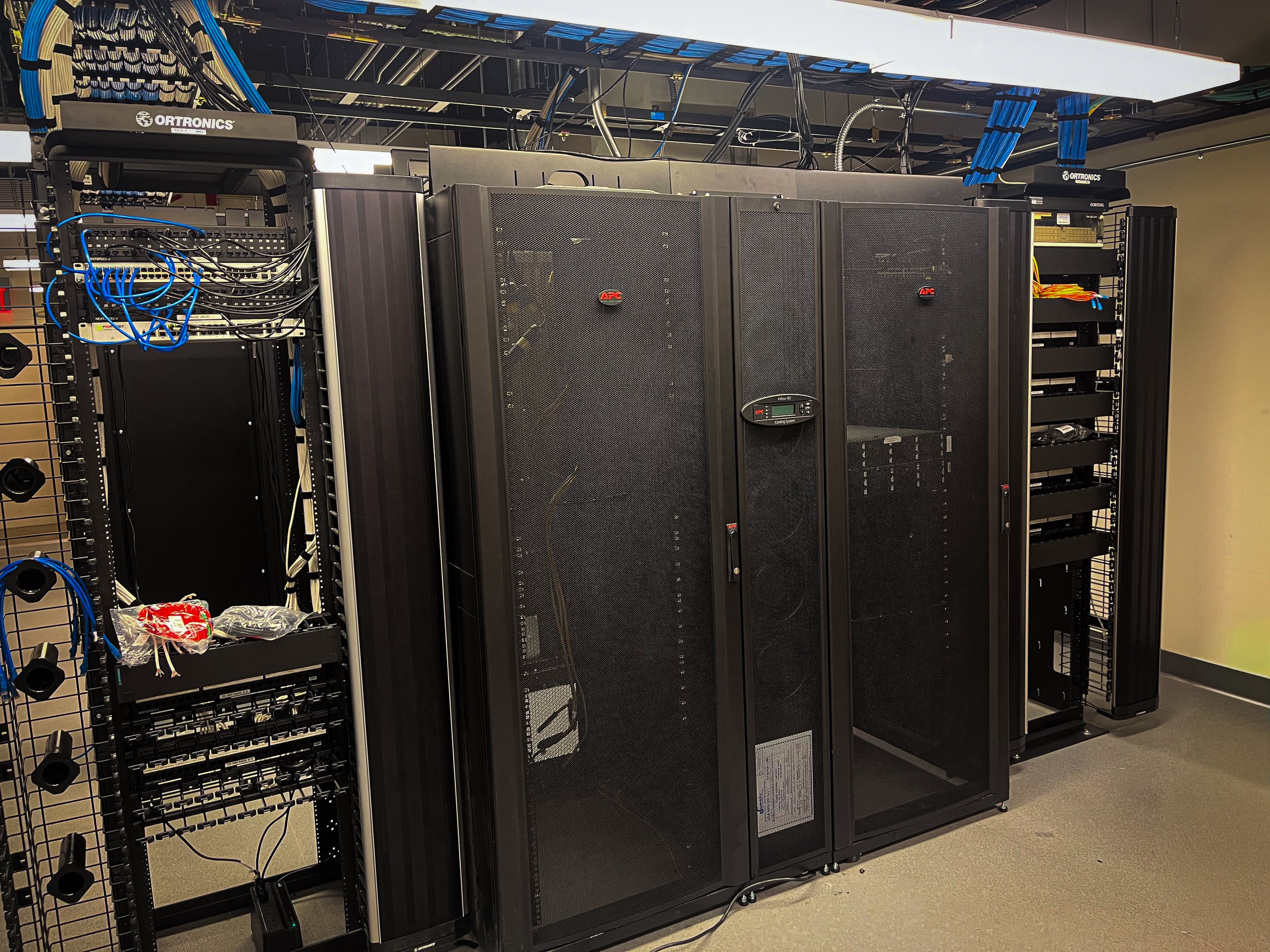 Server room with black server racks and network cables, including a large black cabinet, some open black rack components, and blue cables.