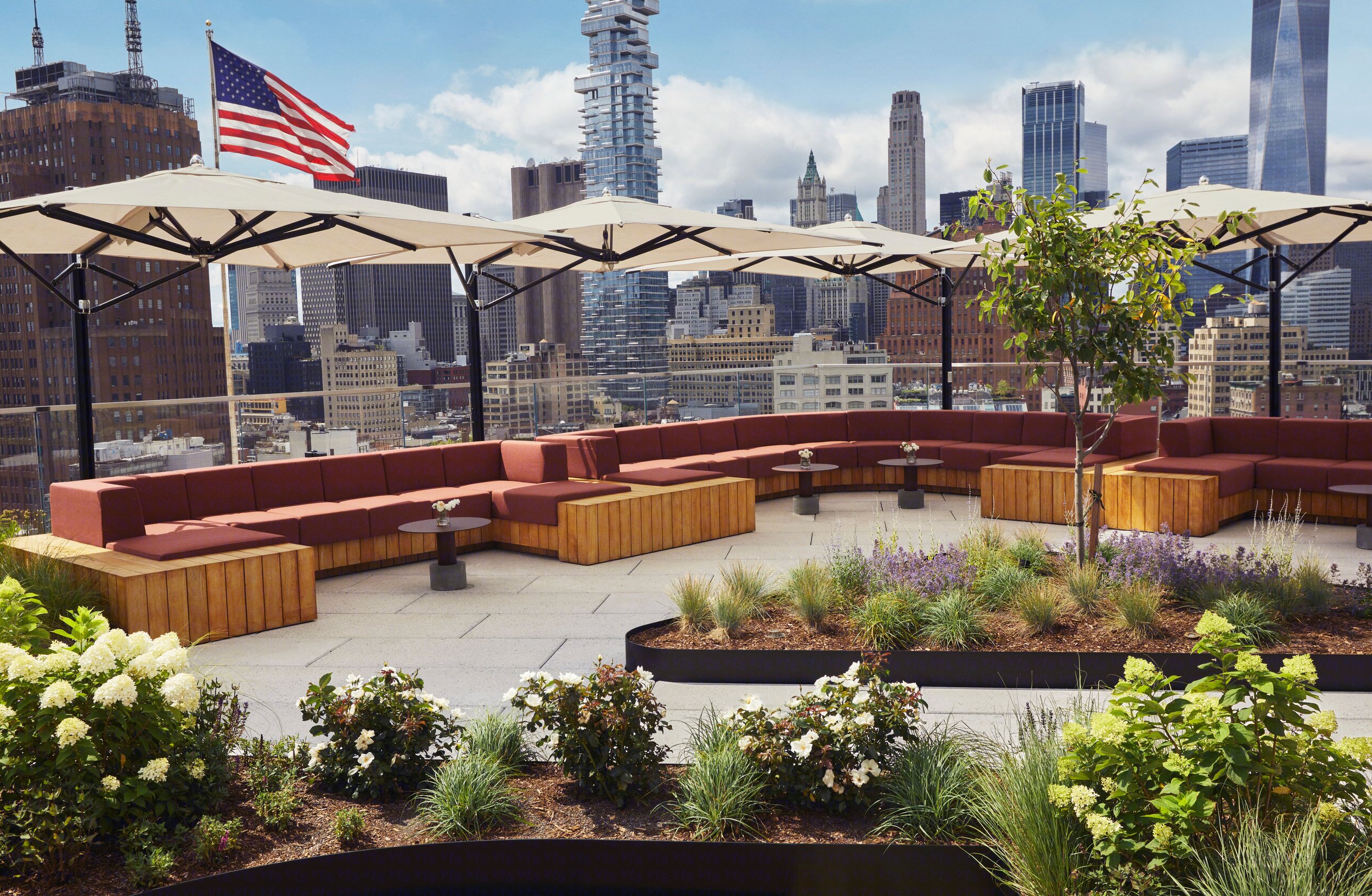 A rooftop terrace with red cushioned seating, umbrellas, a small tree, potted plants, and a city skyline in the background.