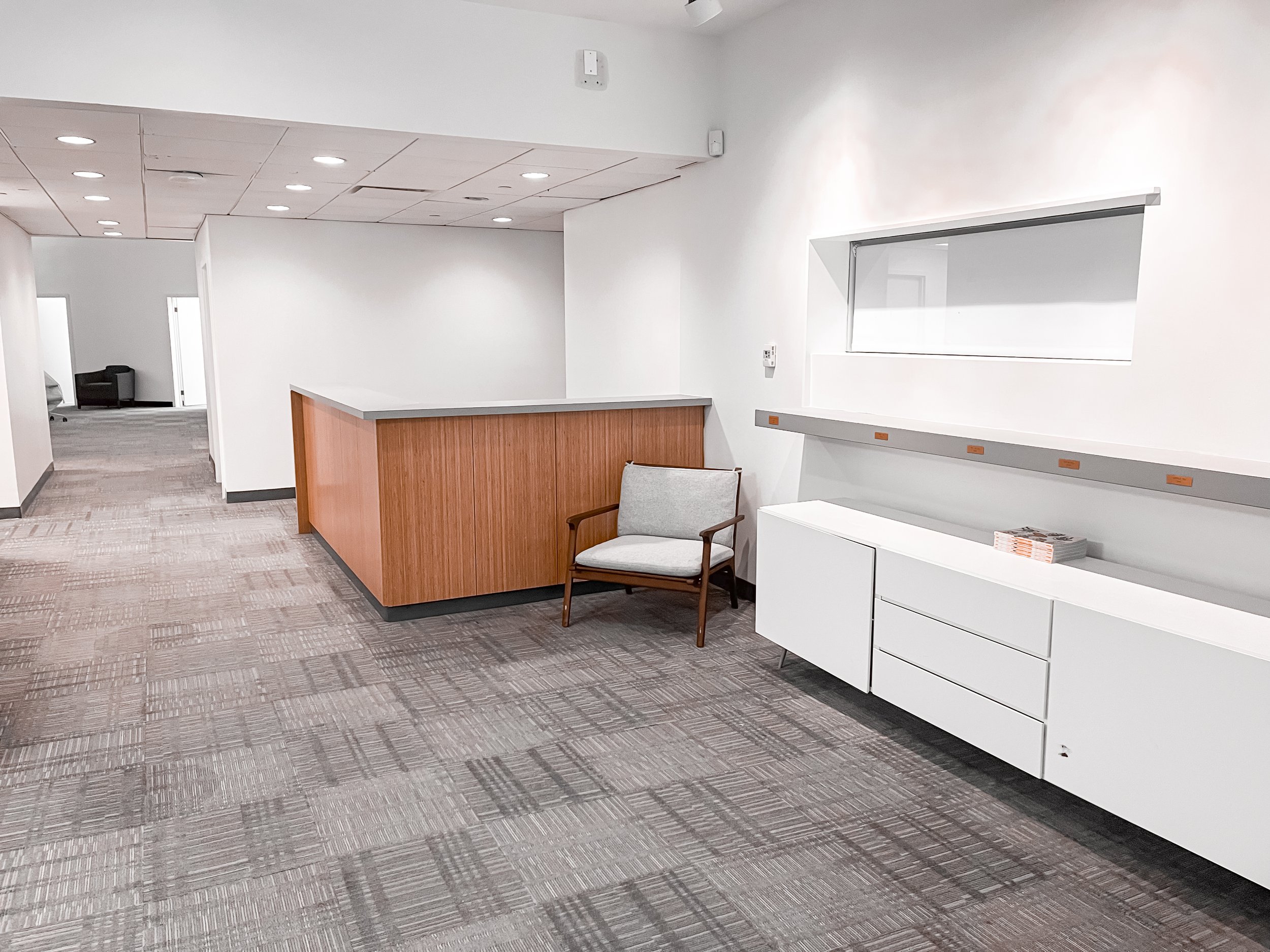 Empty modern office space with a reception counter, a single wooden armchair, and a white cabinet underneath a small horizontal window, with a patterned carpet and white walls.