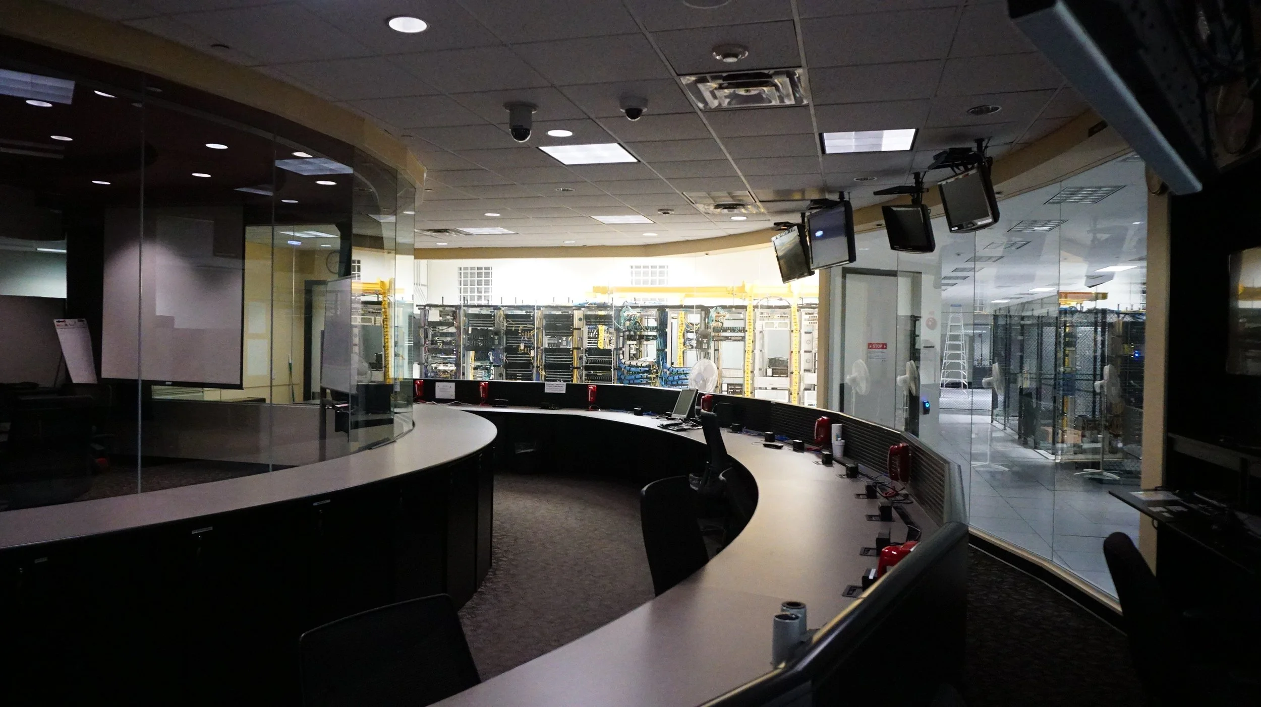 Empty control room with multiple monitors hanging from ceiling, large windows showing server racks outside, and desks with phones and chairs.