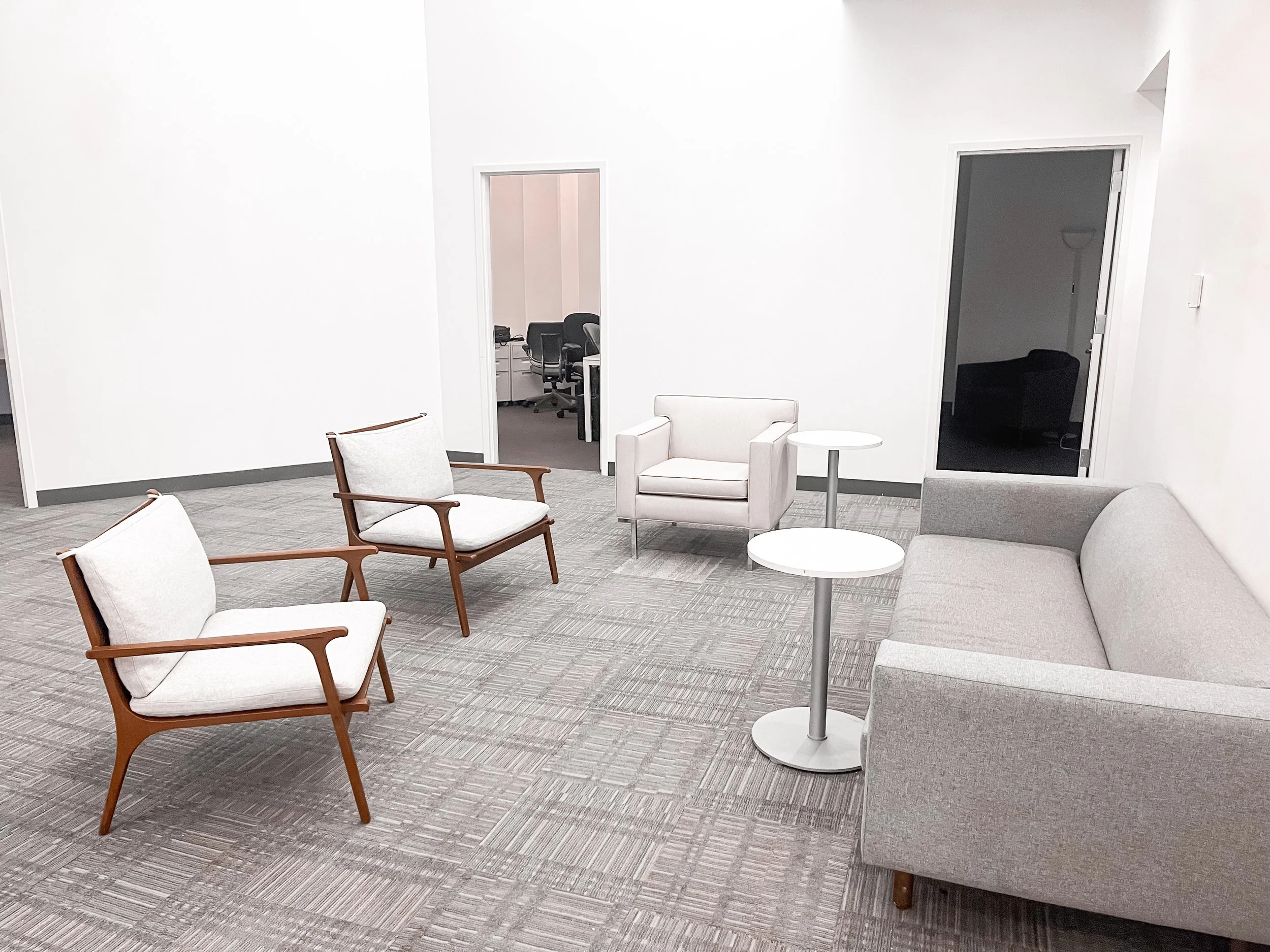 A waiting area with a gray sofa, white armchair, two wooden armchairs with white cushions, and two small white round tables on a patterned carpet, with doorways leading into an office or conference room.
