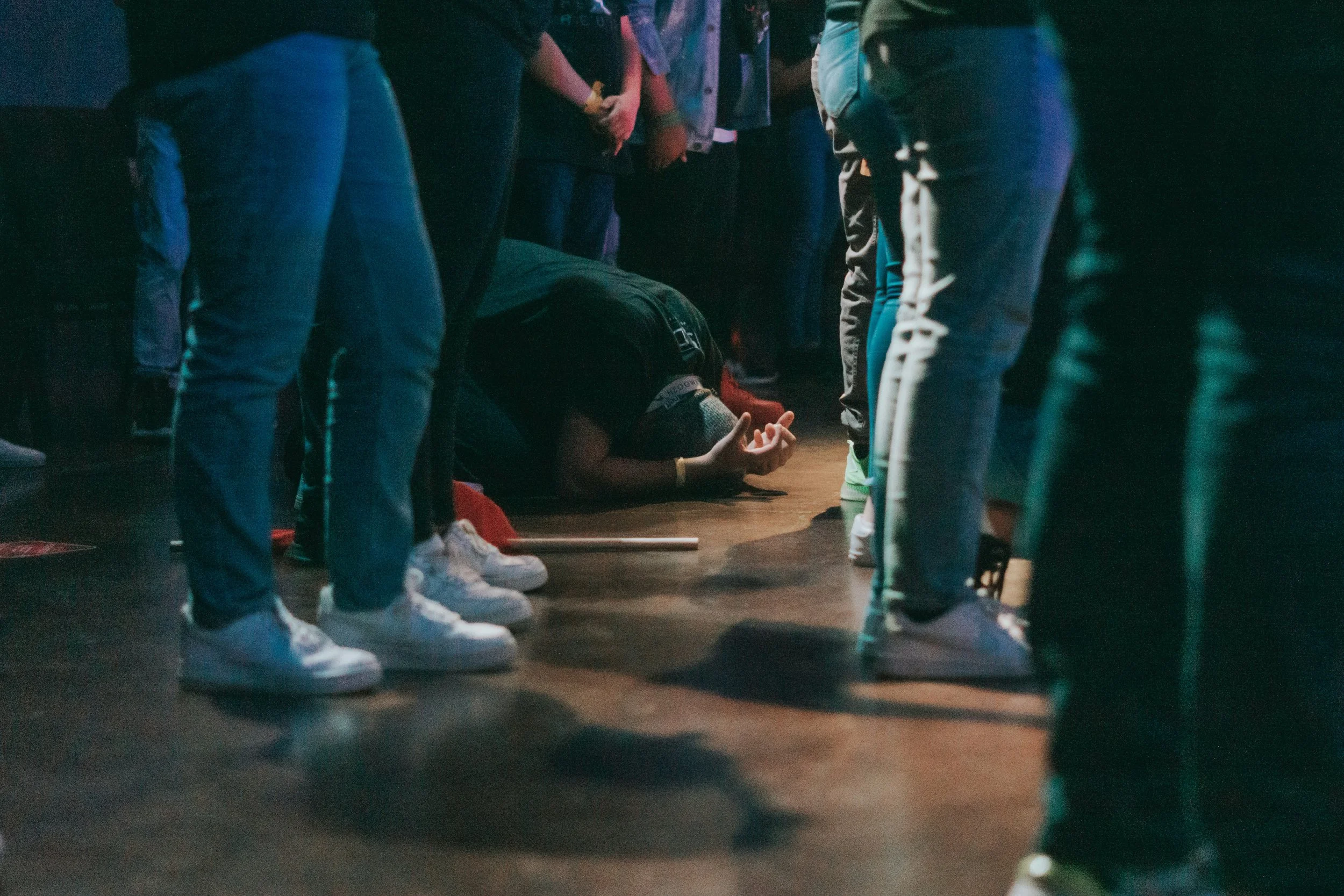Person kneeling, praying, and worshipping on the floor surrounded by standing individuals in a dark setting.