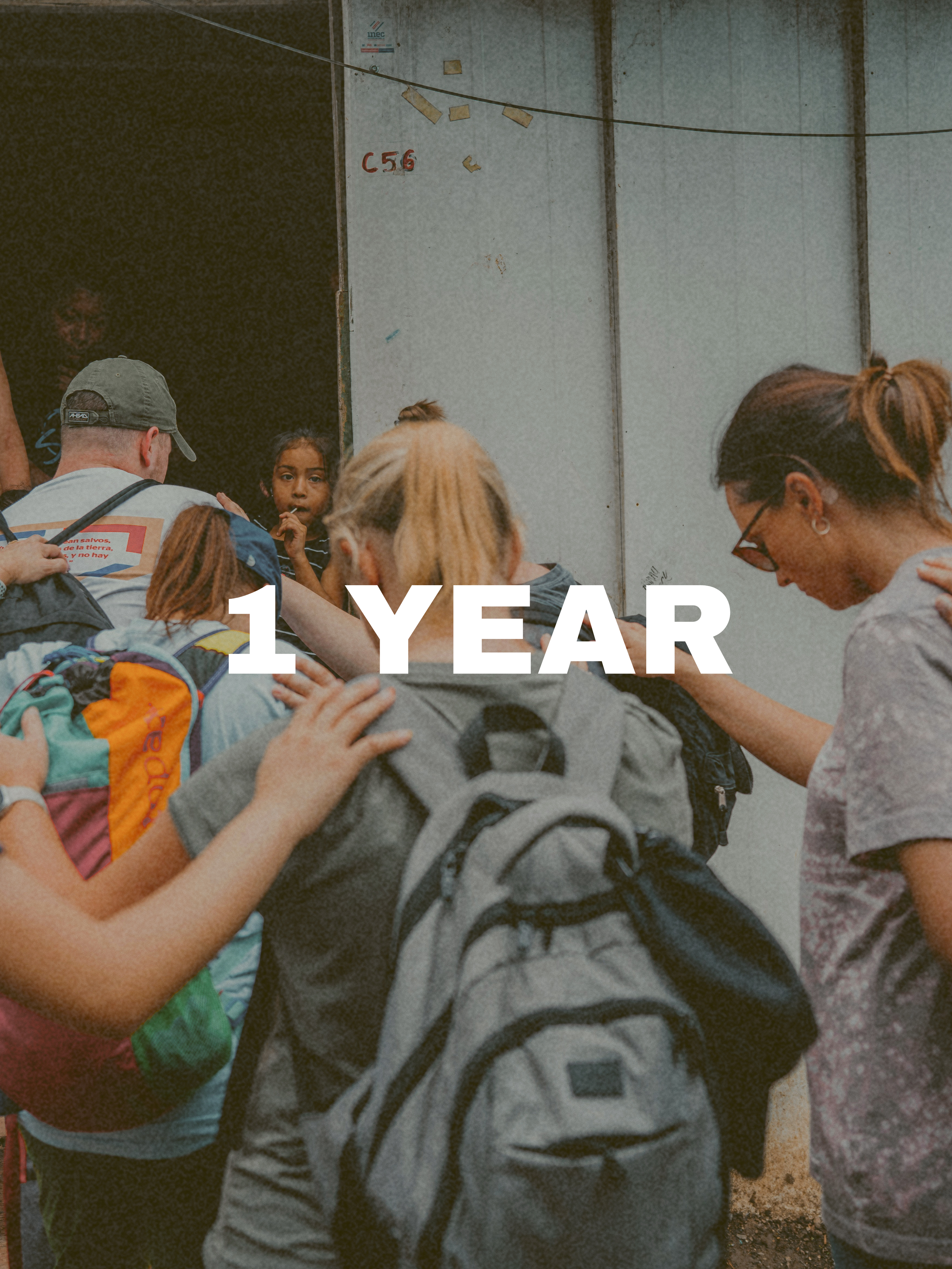 Group of students and adults with backpacks, standing together praying for a woman in a underprivileged community. Text overlay reads "1 YEAR".