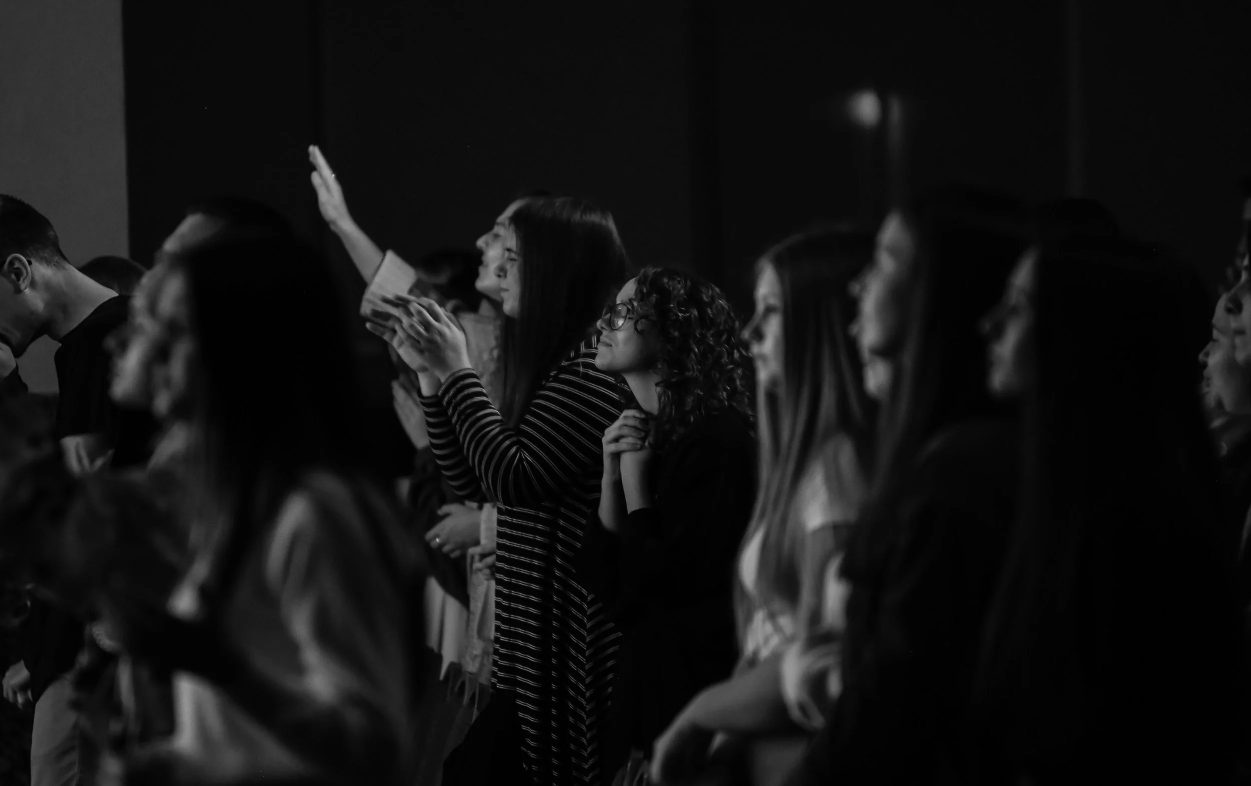 People standing in a dark room, some with eyes closed, one woman with glasses praying with hands clasped, others with raised hands.