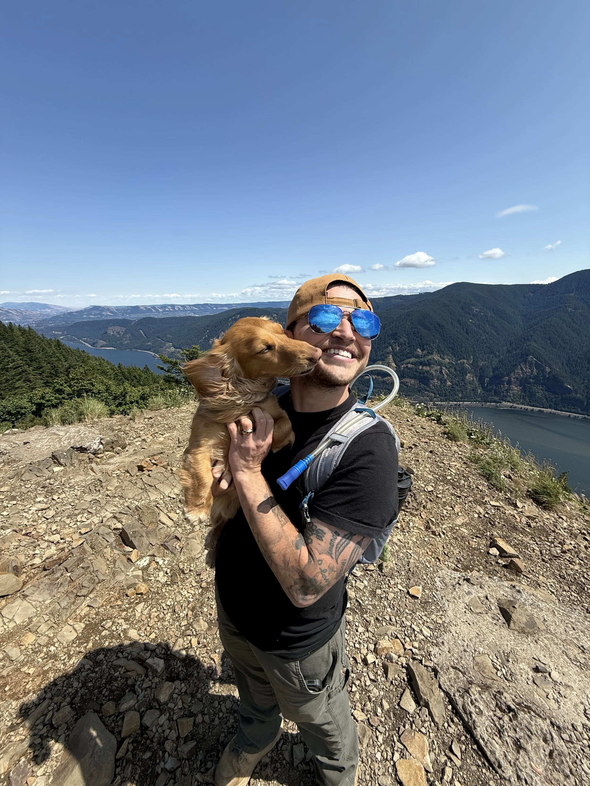 A man with tattoos wearing sunglasses, a tan cap, and a backpack holding a small brown dog on a mountain trail with a scenic view of mountains and a body of water in the background.
