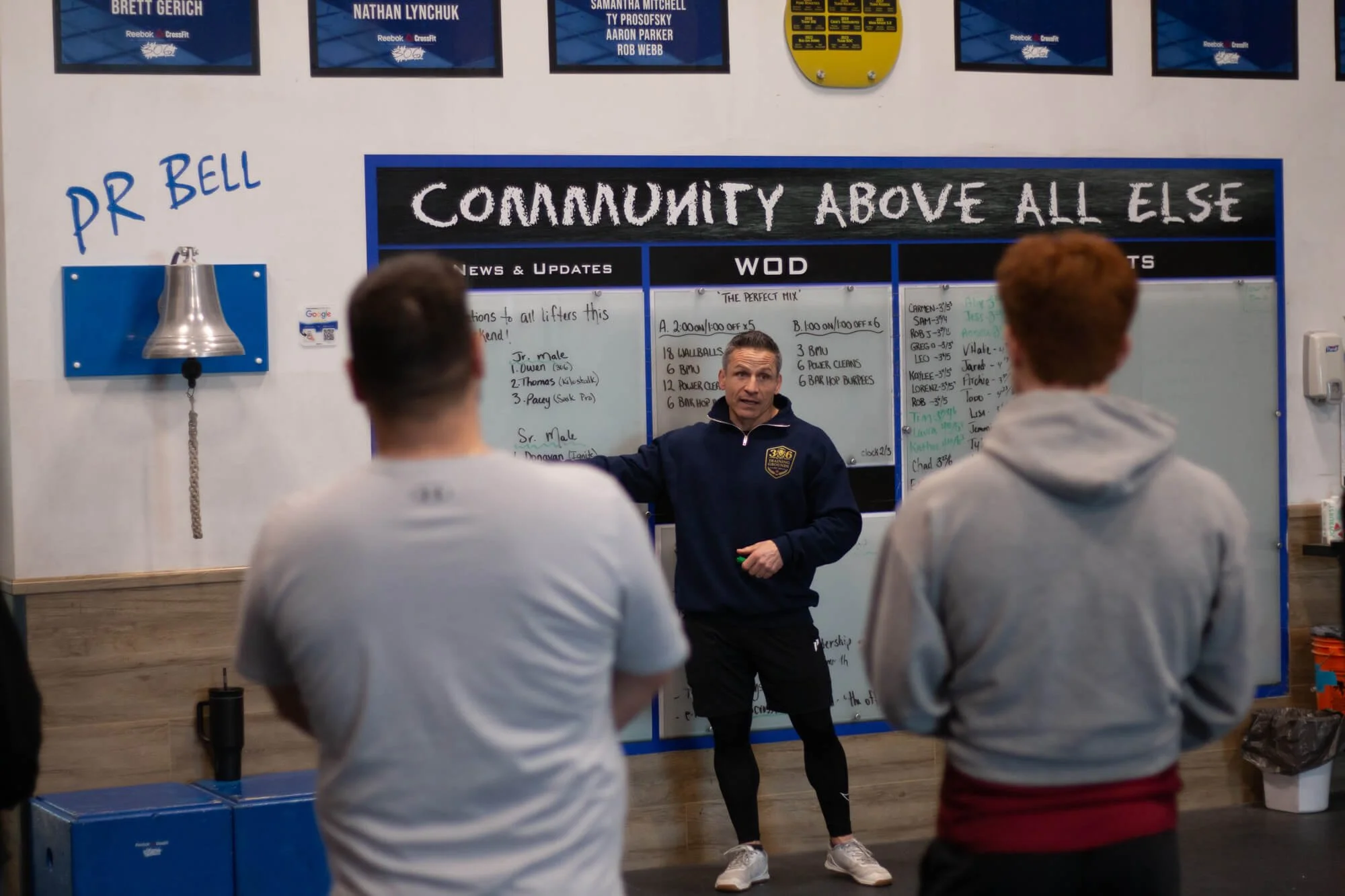 A coach giving instructions to three athletes in a gym, with a whiteboard displaying workout details and a sign reading 'Community Above All Else.'