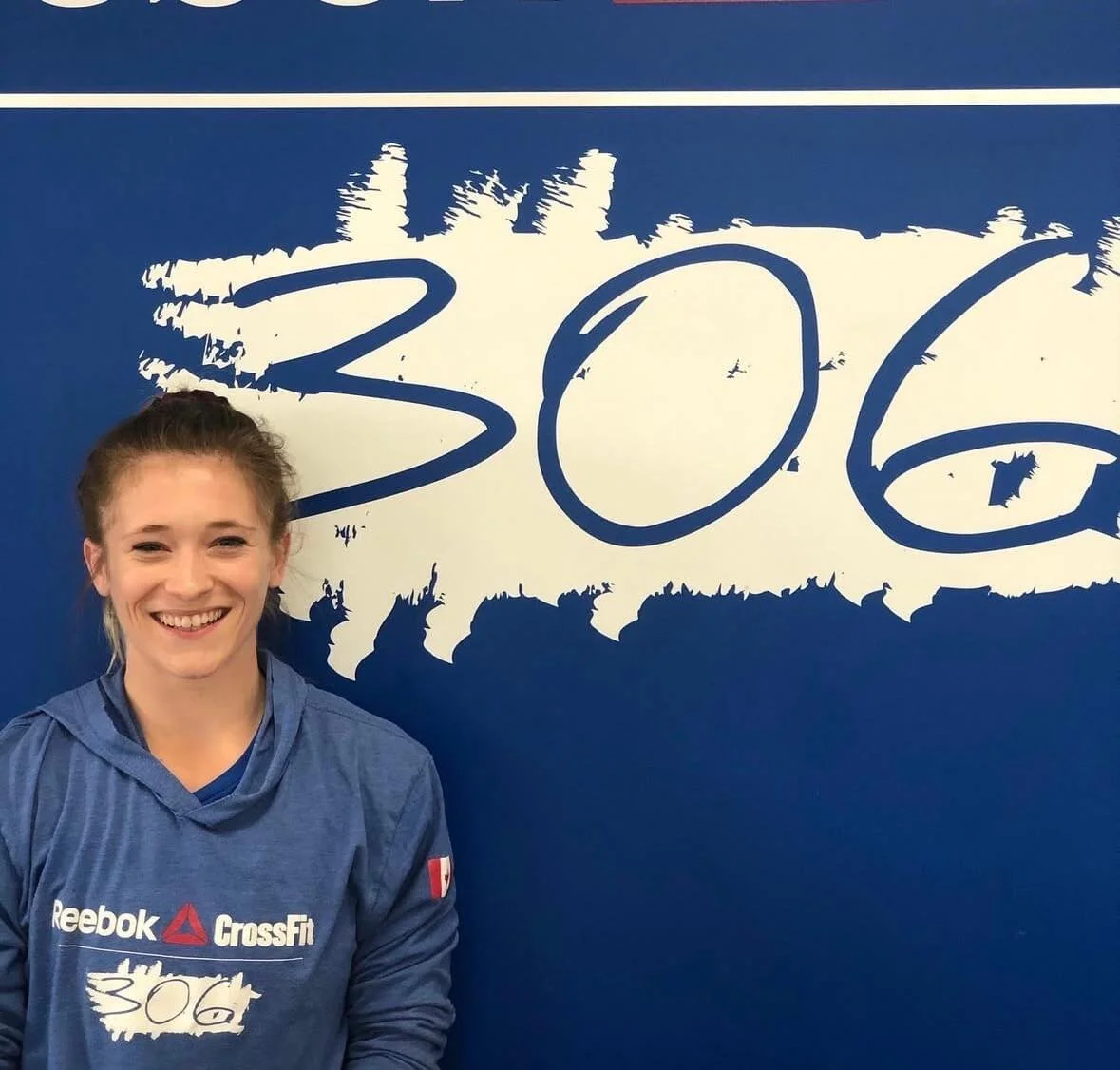 Young woman with a big smile, wearing a blue Reebok CrossFit hoodie, standing in front of a blue wall with a large white graffiti-style '306'.