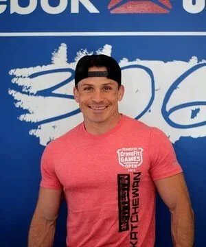 Man in a pink T-shirt and backward cap smiling, standing in front of a blue and white background.