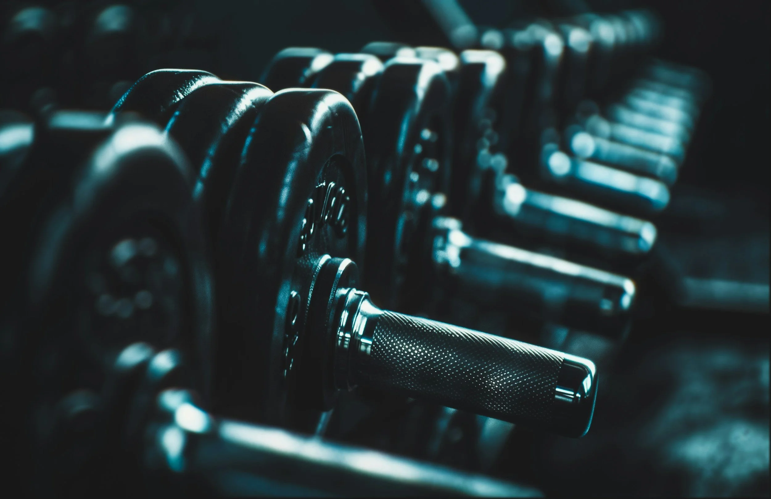 Close-up of black dumbbells arranged in a row in a gym with focus on a textured handle of one dumbbell in the foreground.