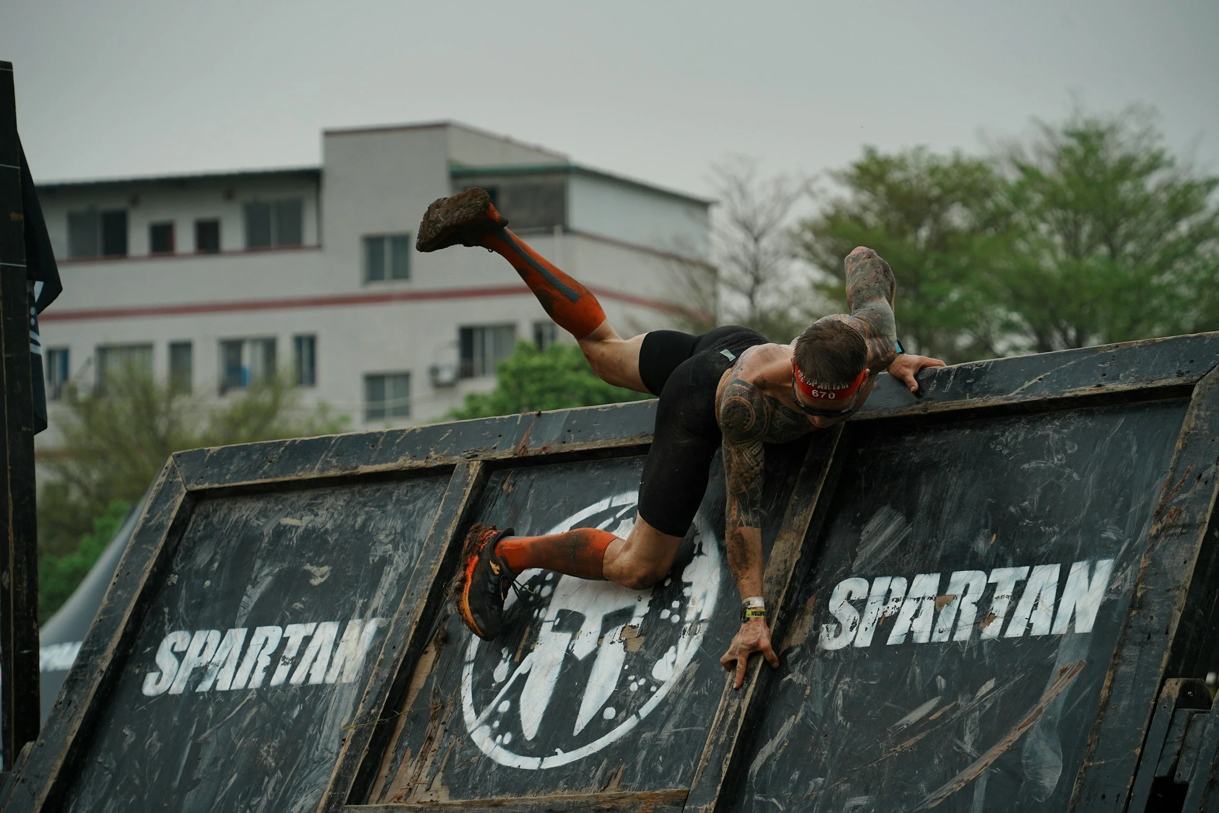 A man with tattoos climbing on a muddy obstacle during a Spartan race, wearing a red headband and black athletic clothing.