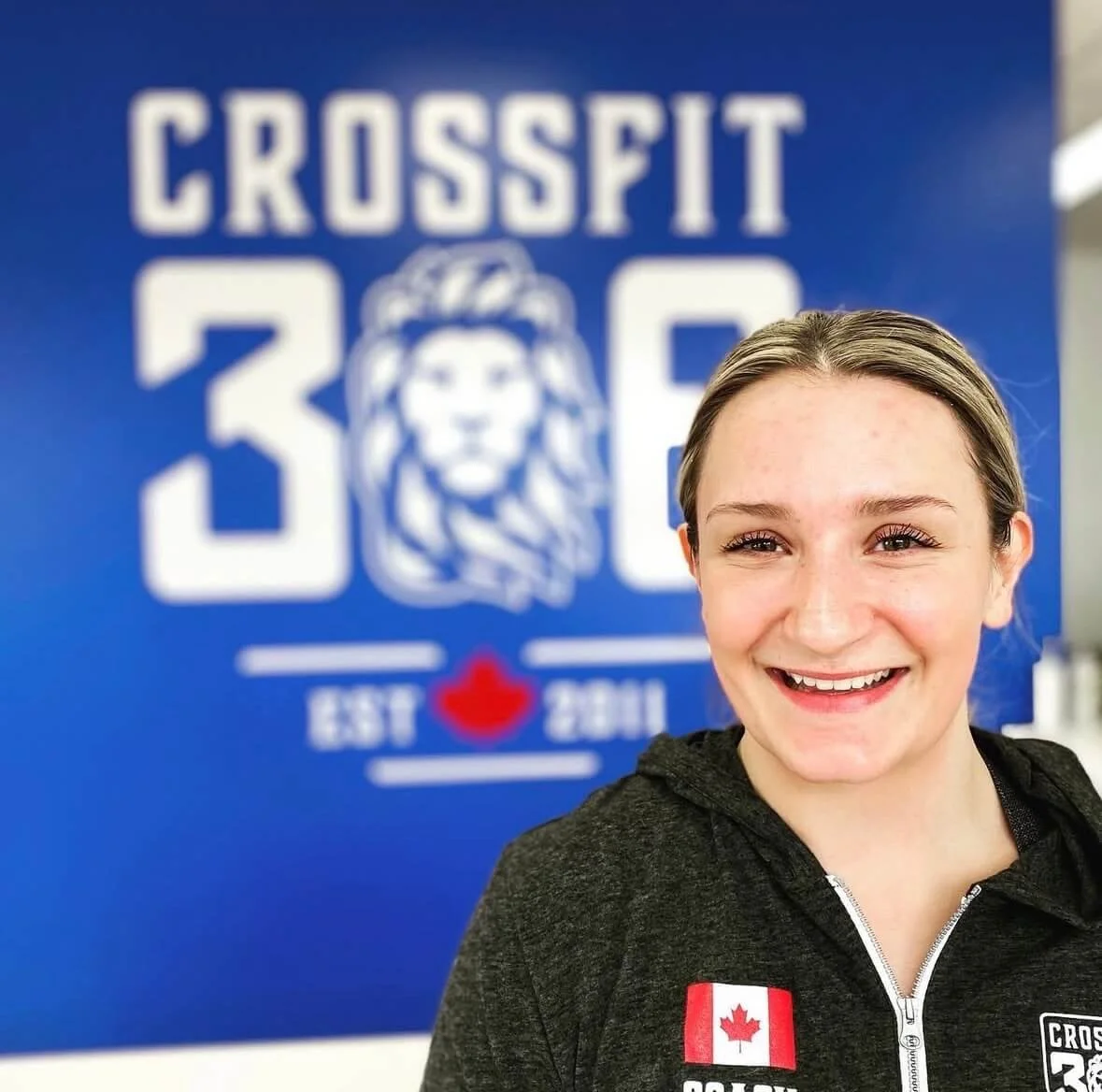 Young woman with a big smile wearing a dark zip-up hoodie with a Canadian flag patch, standing in front of a blue CrossFit gym sign featuring a lion logo and the text "CROSSFIT 3D EST 2011."