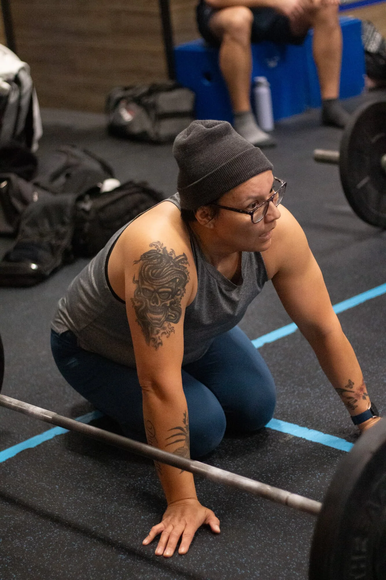 A woman with tattoos, wearing glasses, a gray tank top, and a black beanie, is kneeling on the gym floor preparing to lift a barbell.
