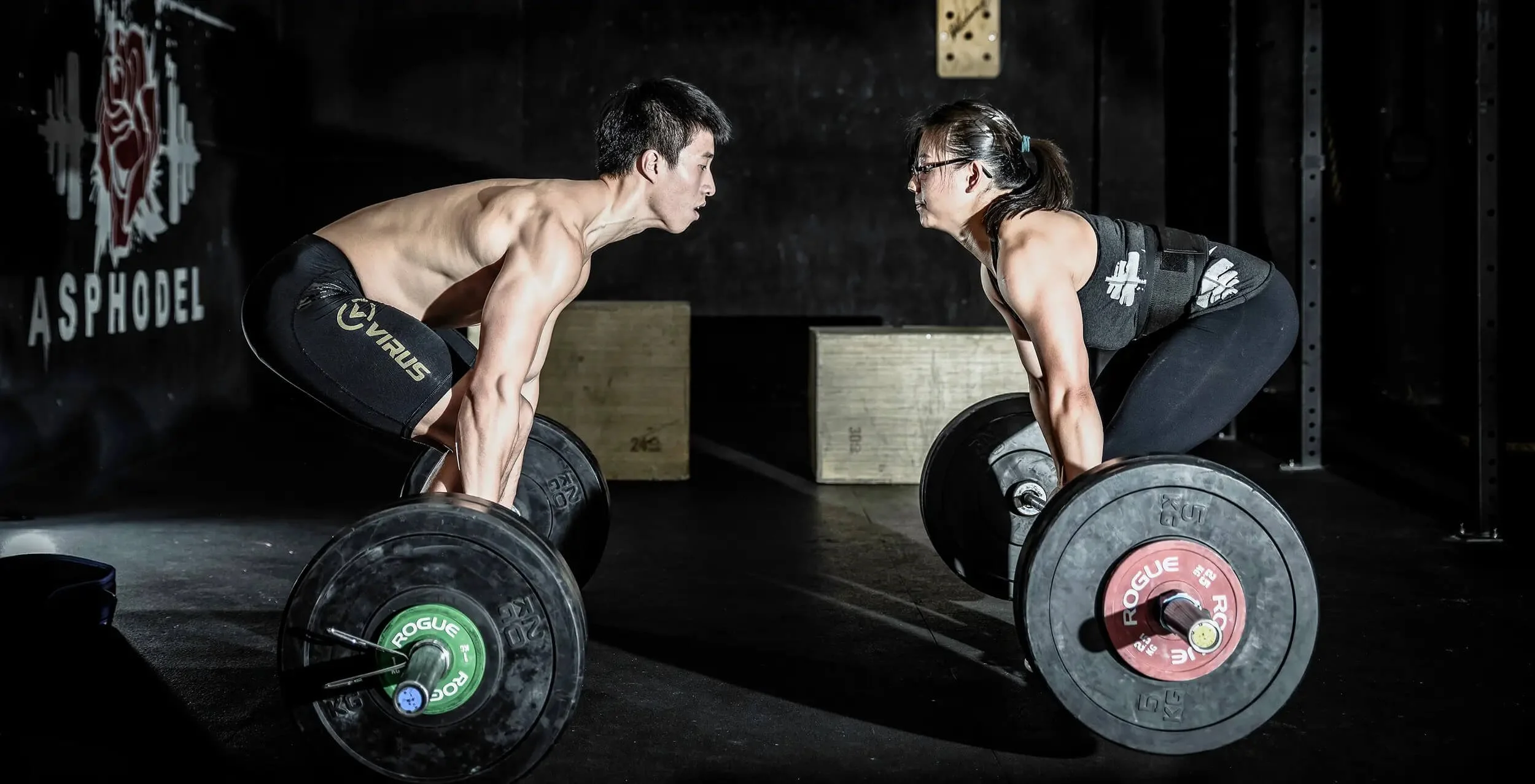 A man and a woman in a gym, preparing to lift barbells during a deadlift exercise, facing each other on a dark gym floor.