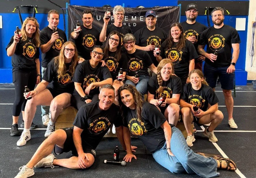 Group of people, wearing black T-shirts with a lion logo and text, posing together inside a gym, holding drinks, smiling for the camera.