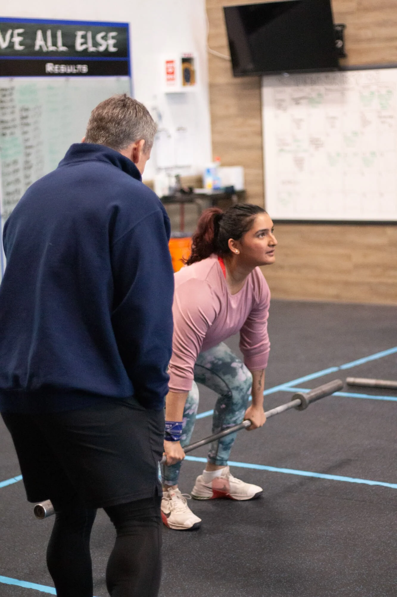 A woman in a pink top and patterned leggings lifting a barbell in a gym while a man in a blue jacket watches her.