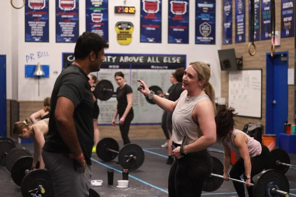 A woman in a gym talking to a man, with others lifting weights in the background.