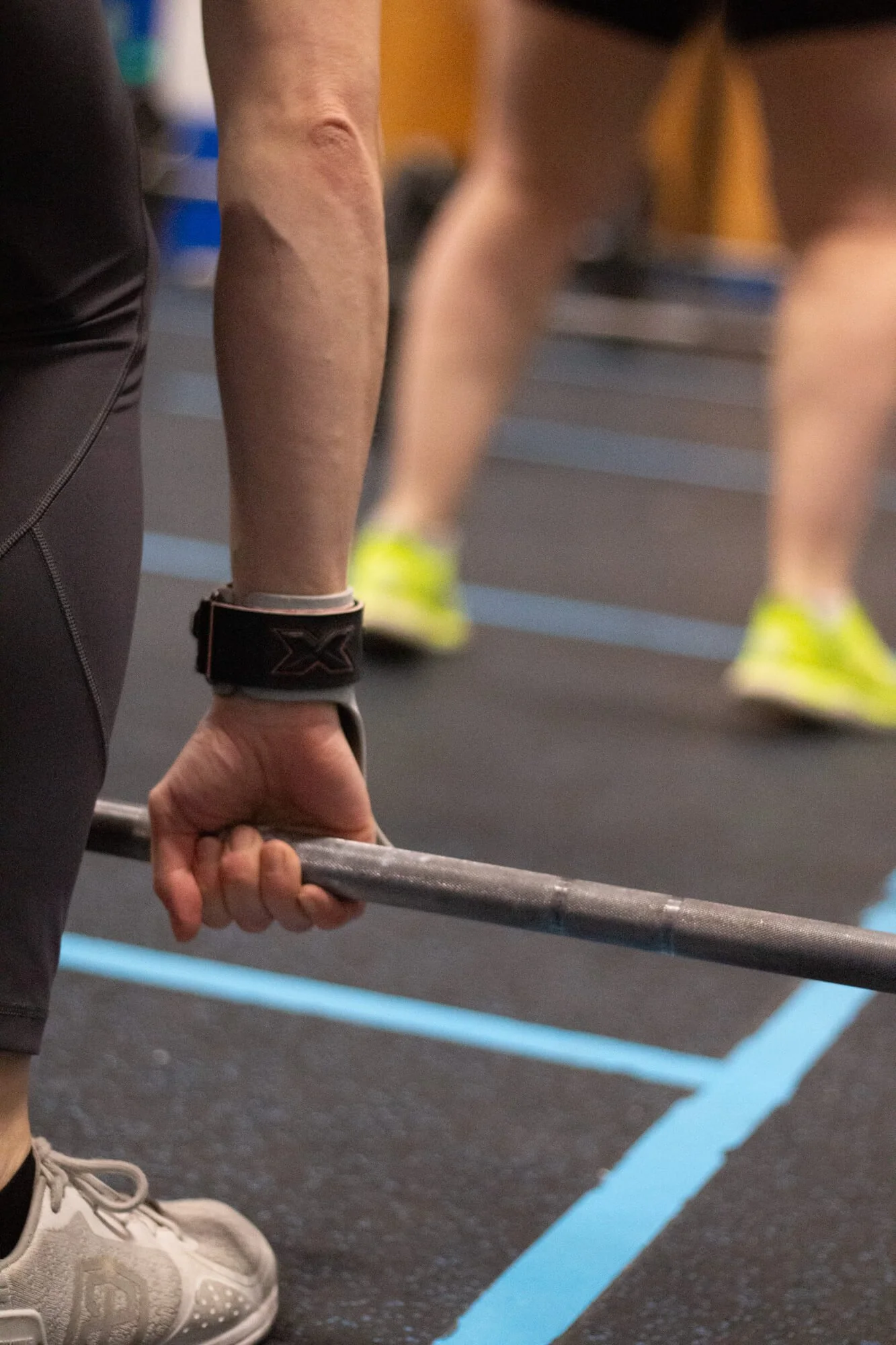 Close-up of a person gripping a barbell during a workout in a gym, with other individuals in athletic wear visible in the background.