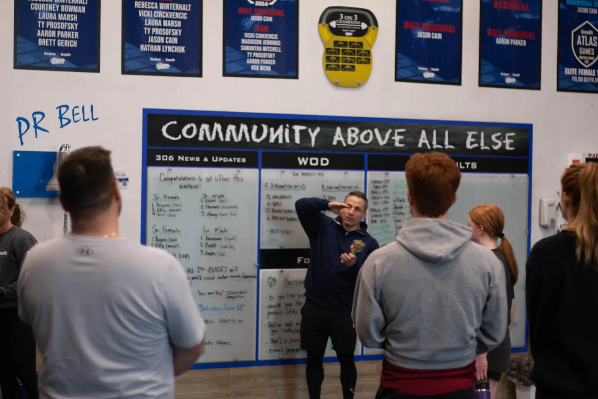 A police officer giving a speech or instructions to a group of young people inside a gym or training facility. The group is standing in front of a large community bulletin board with various posters and information.