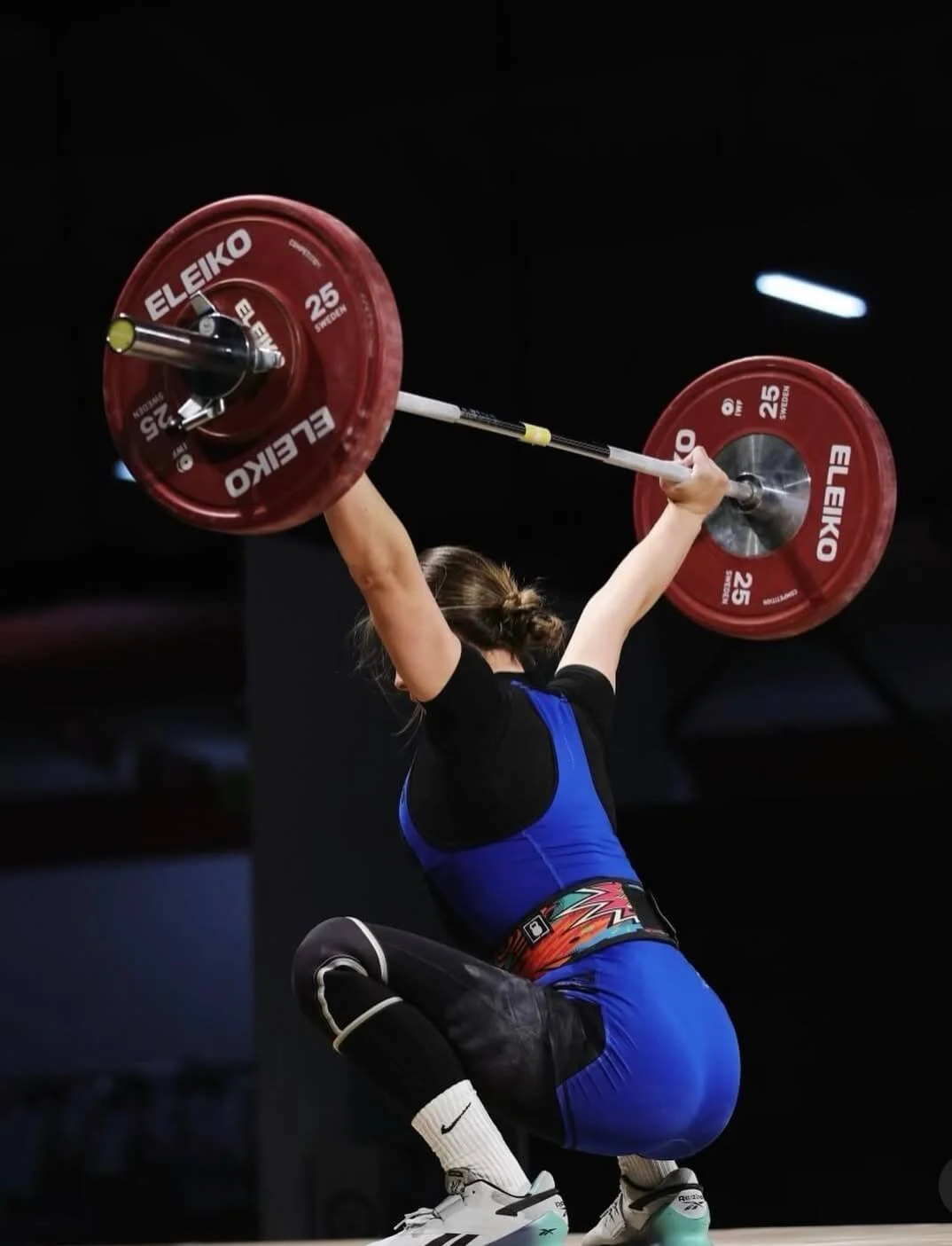 A woman squats and lifts a barbell overhead during weightlifting, wearing a blue and black athletic suit with knee-high socks.