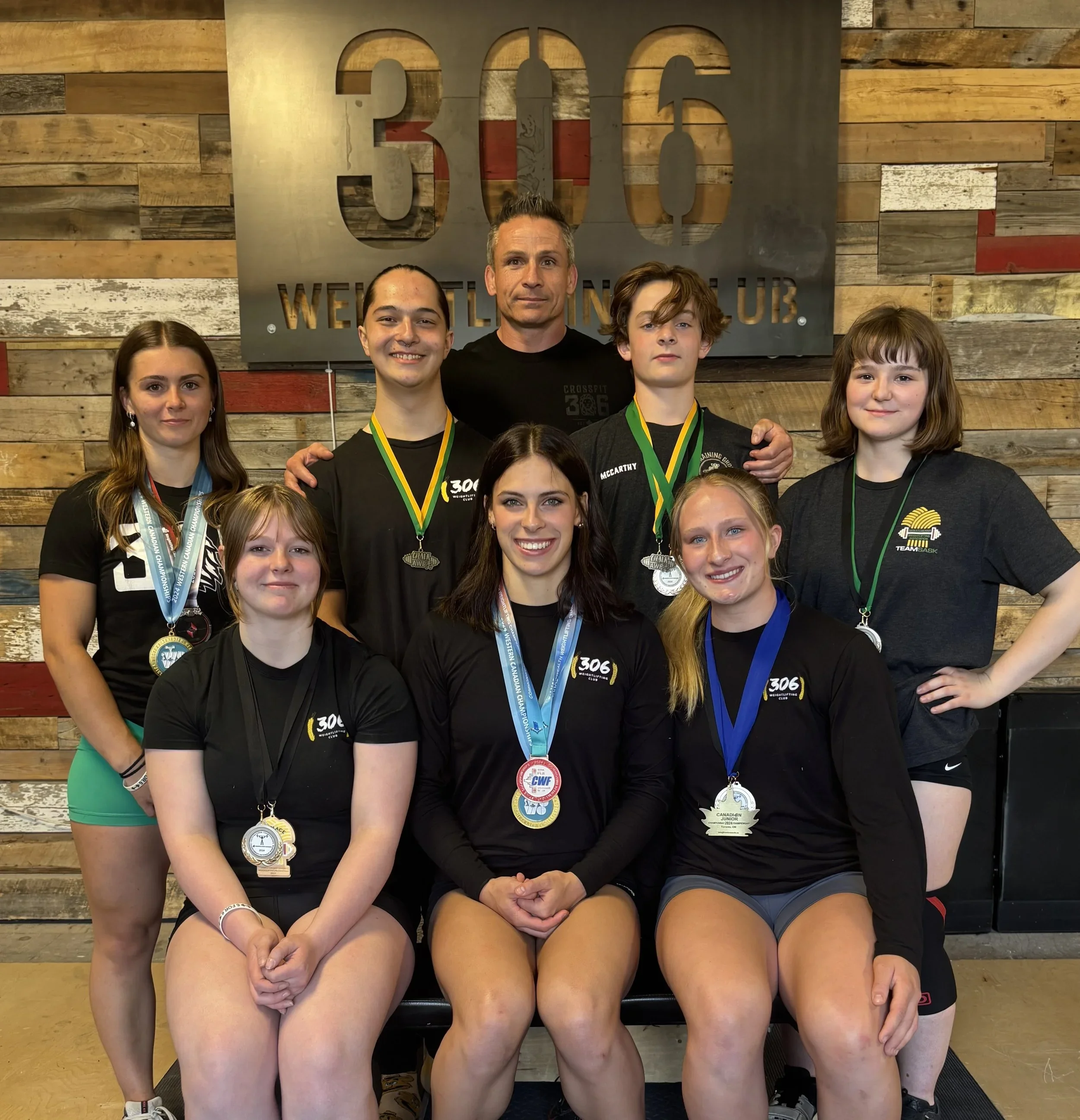 Group of young athletes with medals and a coach at a weightlifting club called 306 Weightlifting Club, standing in front of a wooden wall and a sign that reads 306 Weightlifting Club.