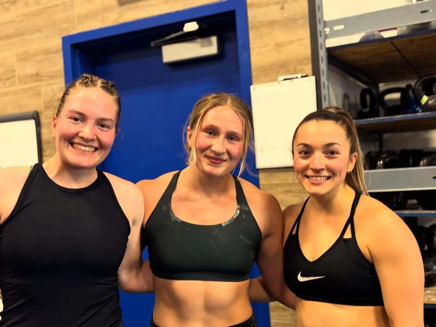 Three young women in athletic wear smiling and posing together in a gym or fitness studio.