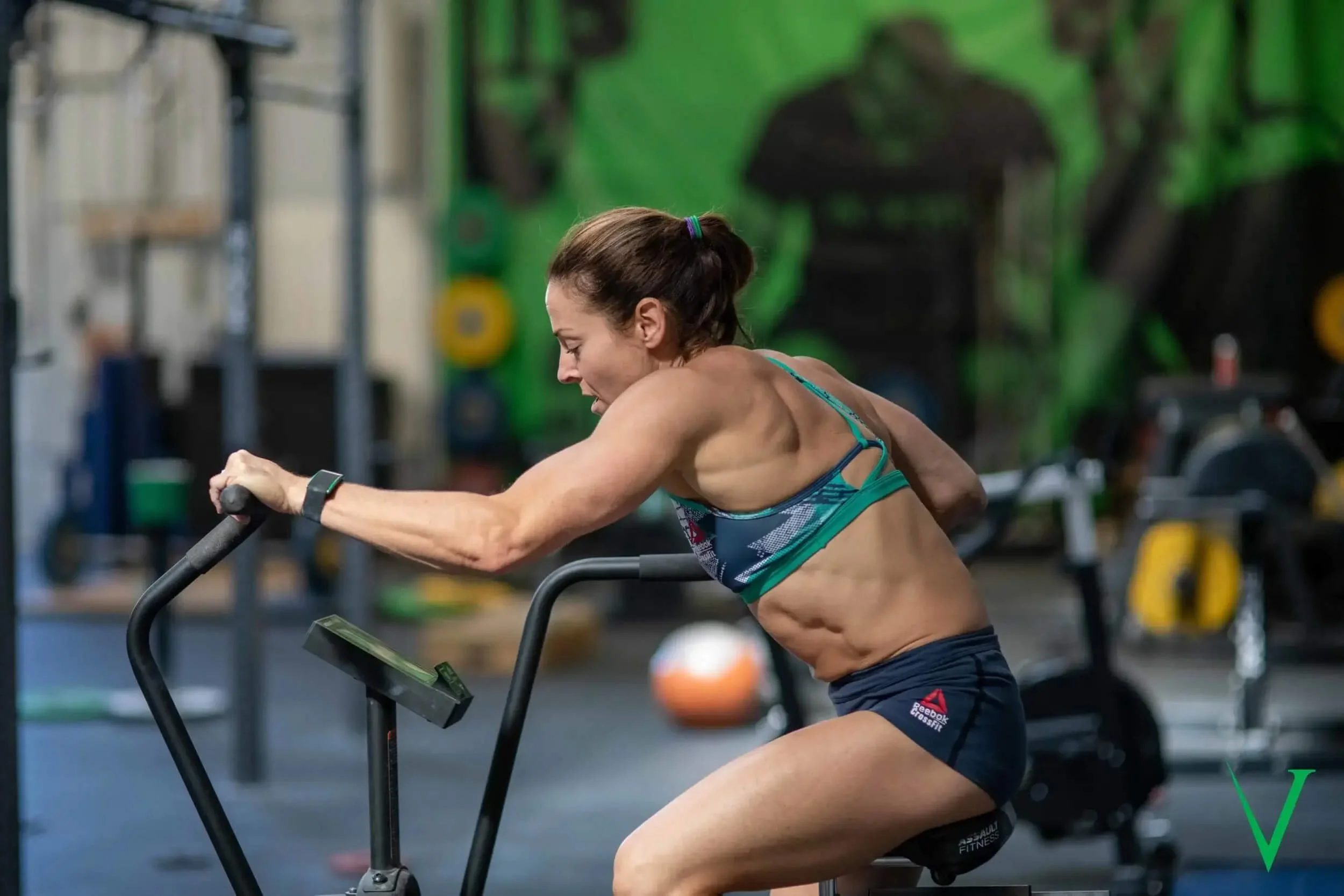 A woman with muscular arms and abs using a stationary exercise bike in a gym.
